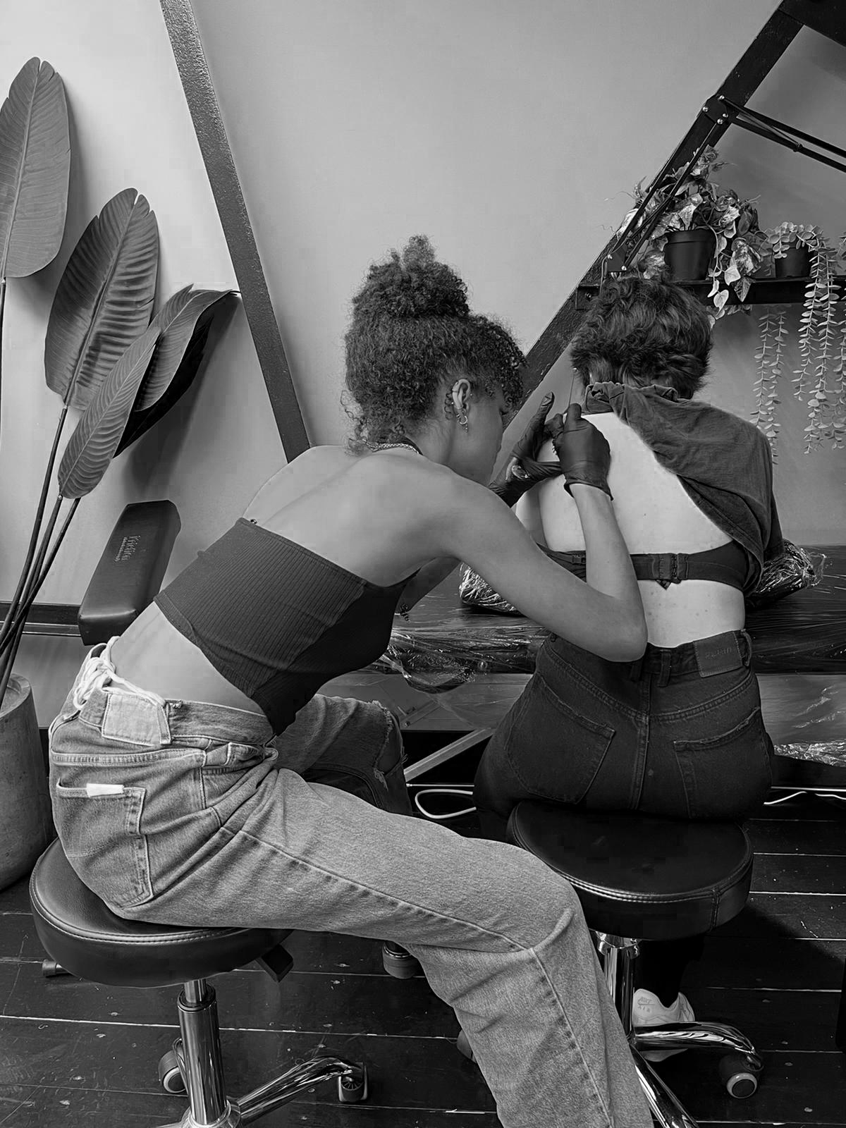 Two women at a tattoo studio, one tattooing the other's lower back.