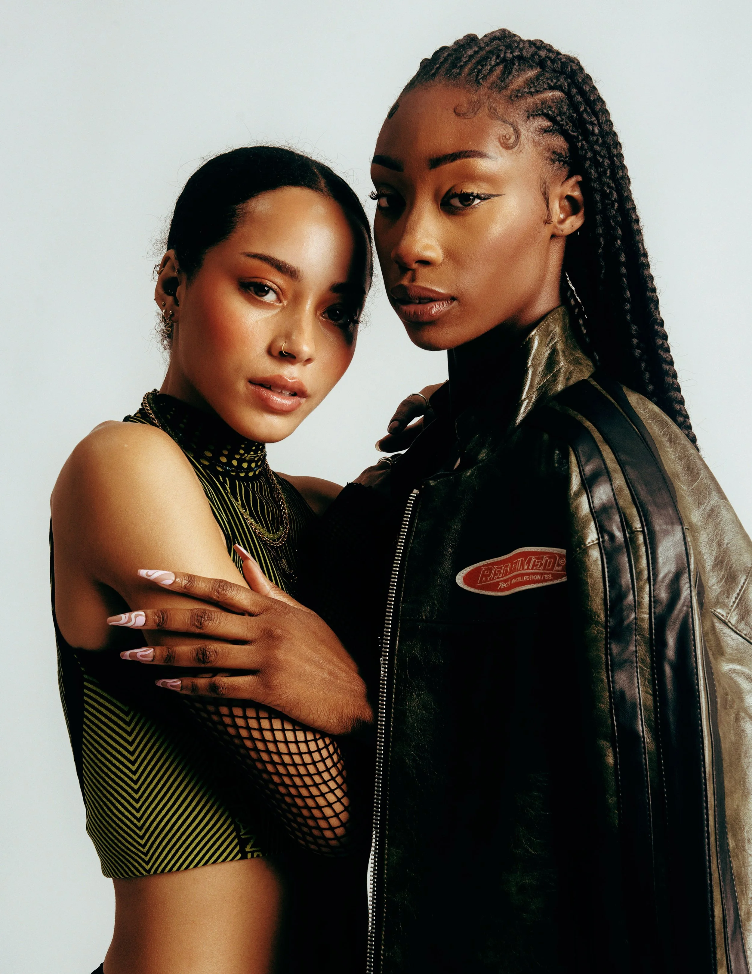 Two women with dark skin and long braided hair are posed closely together against a plain white background. One woman wears a black and yellow patterned sleeveless top with fishnet sleeves, while the other wears a black leather jacket with a patch on