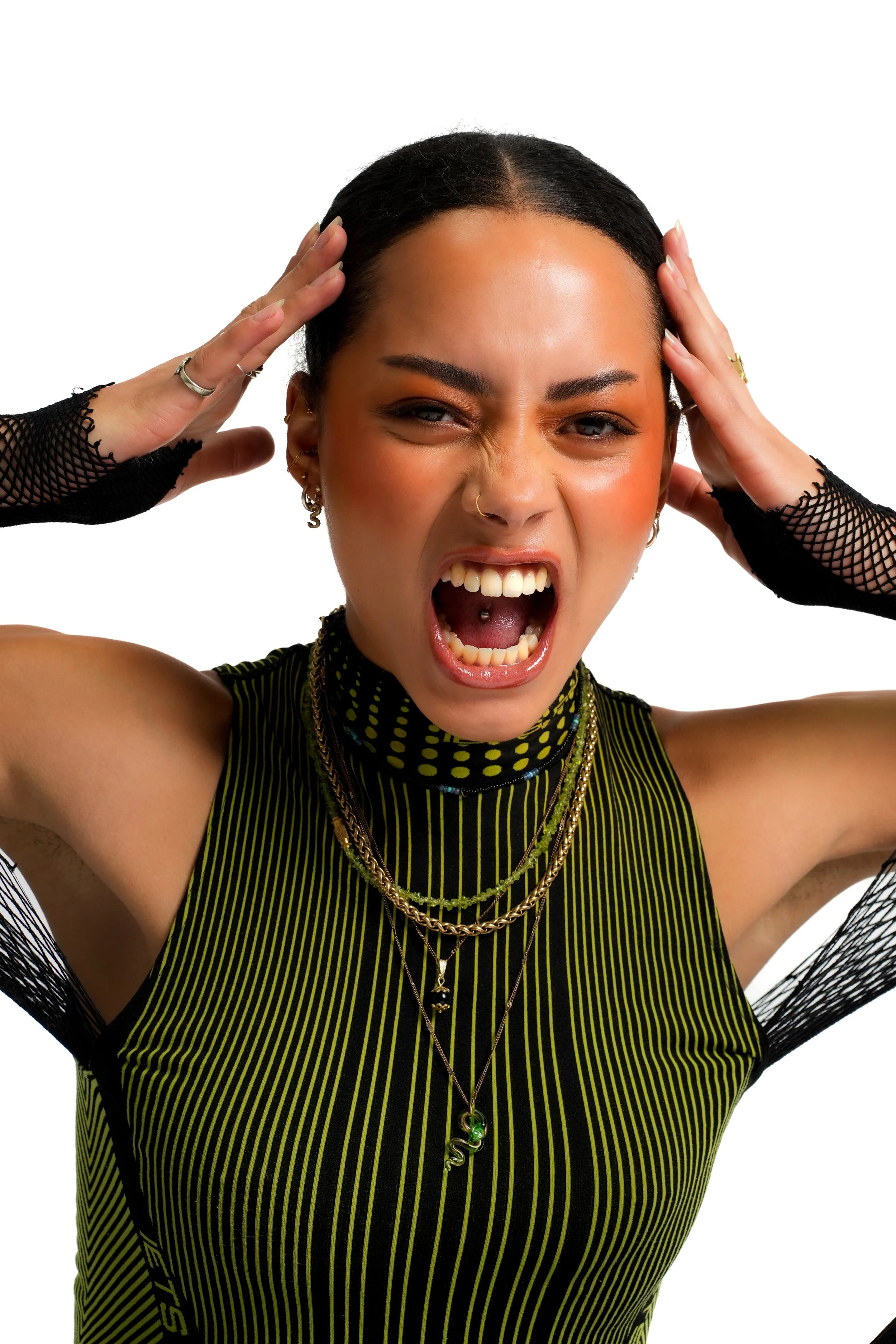 A woman with black hair, tattoos, and jewelry, wearing a black and yellow striped top and black mesh arm sleeves, makes an angry expression with her mouth open and hands on her head.