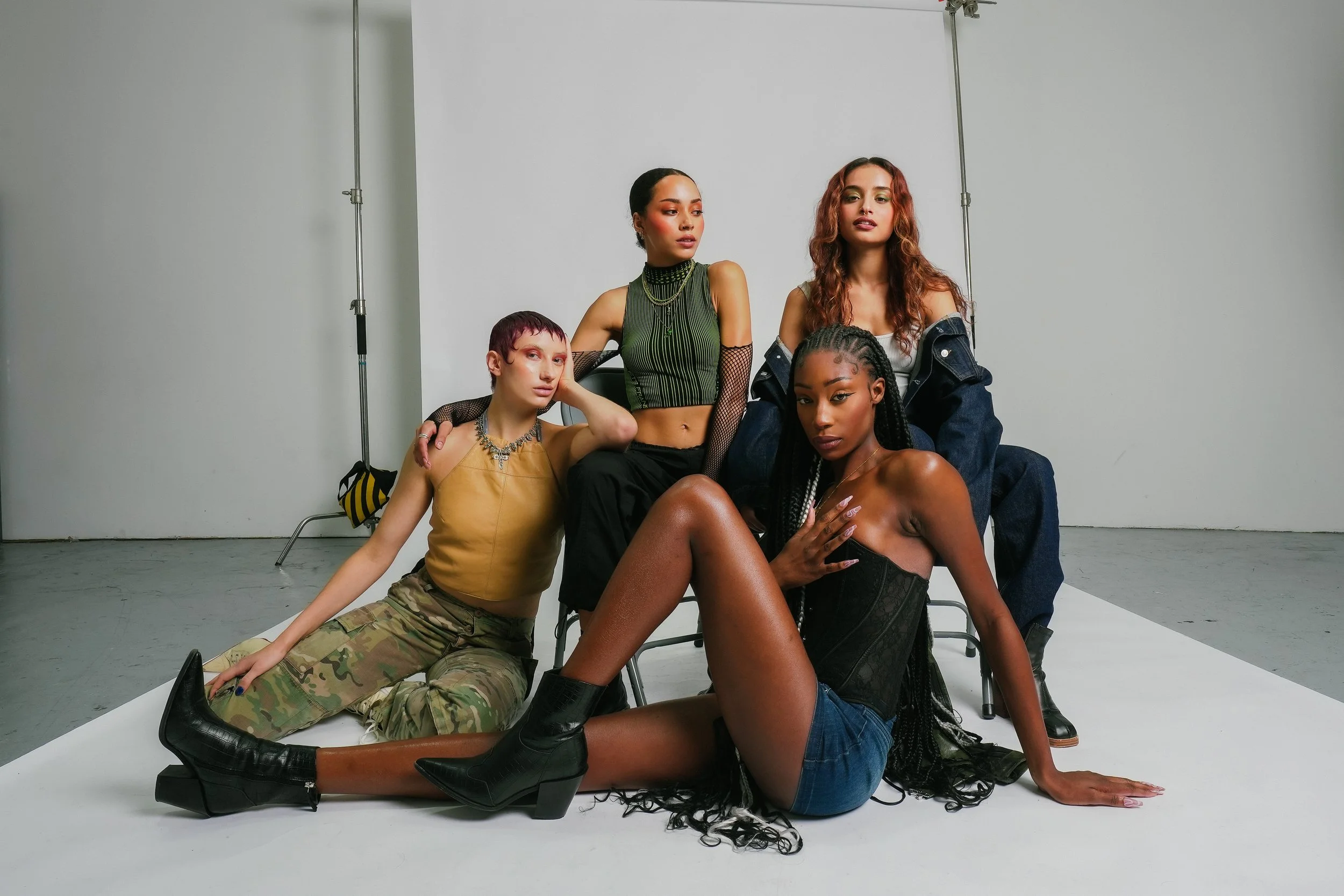 Group of five diverse women posing together against a plain studio background, some sitting and some standing, showcasing fashion and confident expressions.