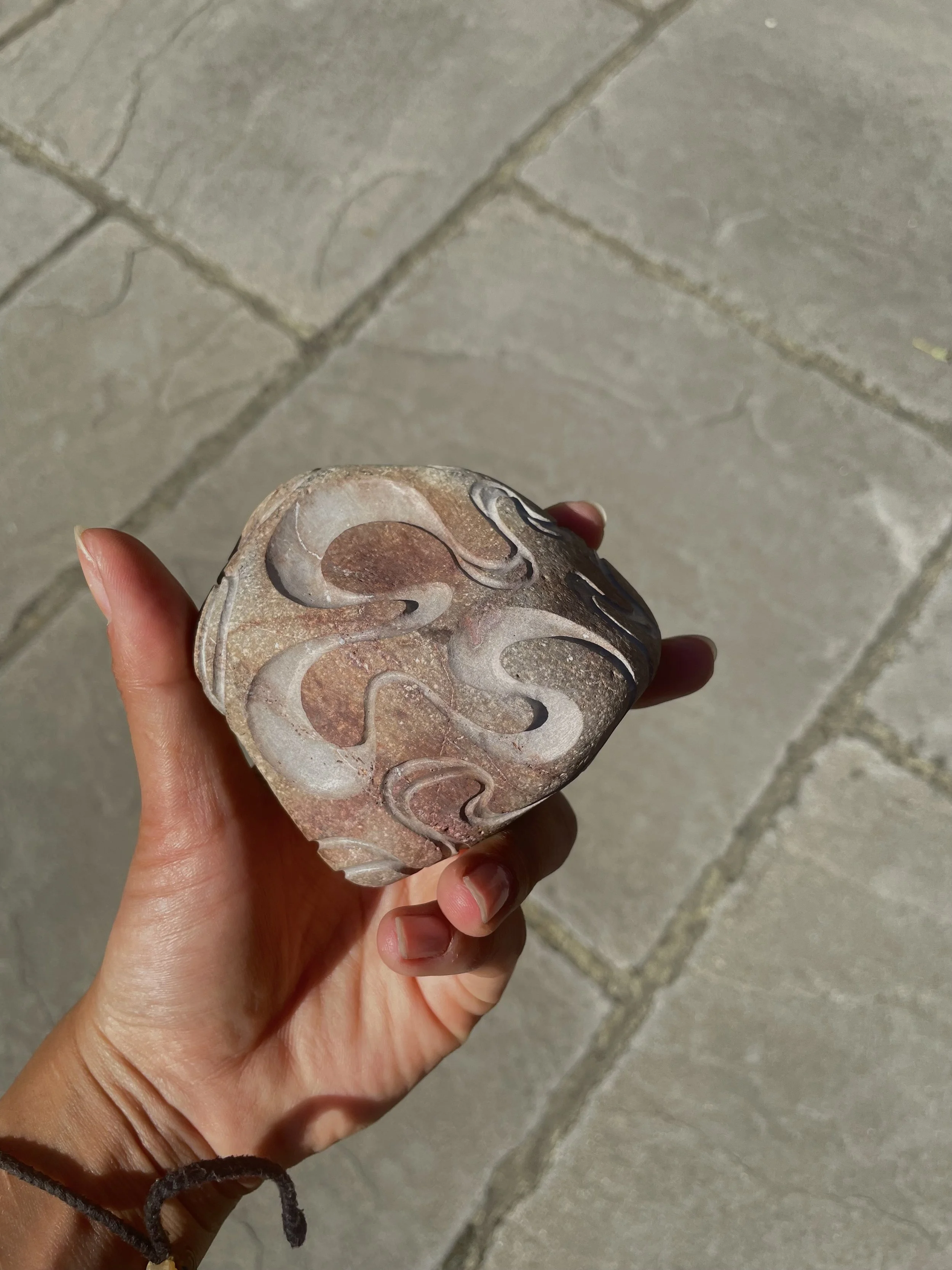 Hand holding a round stone with abstract swirl patterns, against a tiled floor background.
