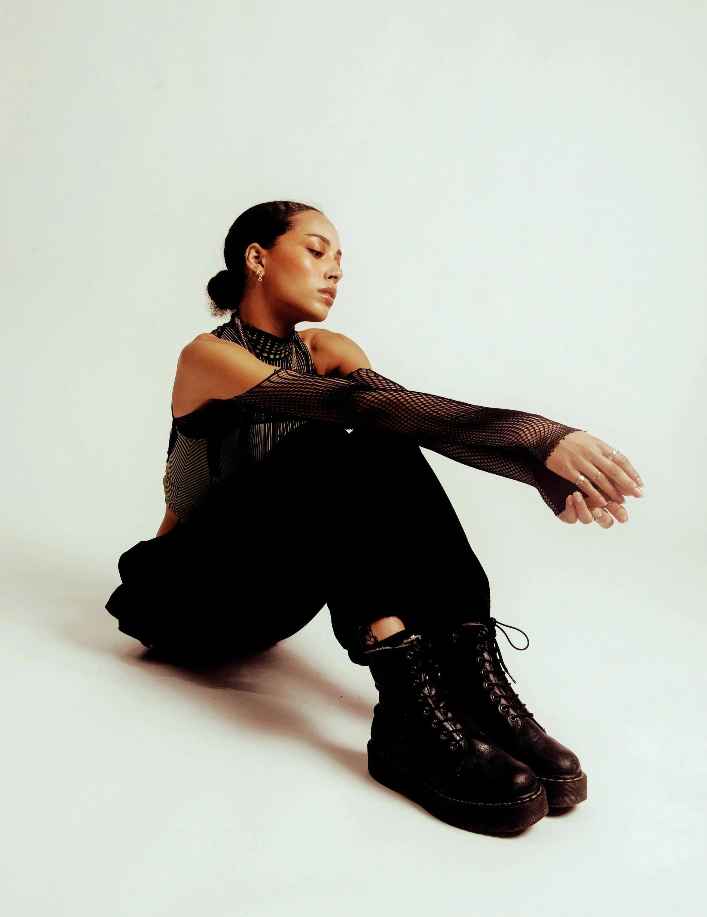 A woman with dark hair tied back, wearing a black fishnet top, black pants, and black lace-up boots, sitting on the floor with her knees bent and arms resting on her knees, against a plain white background.