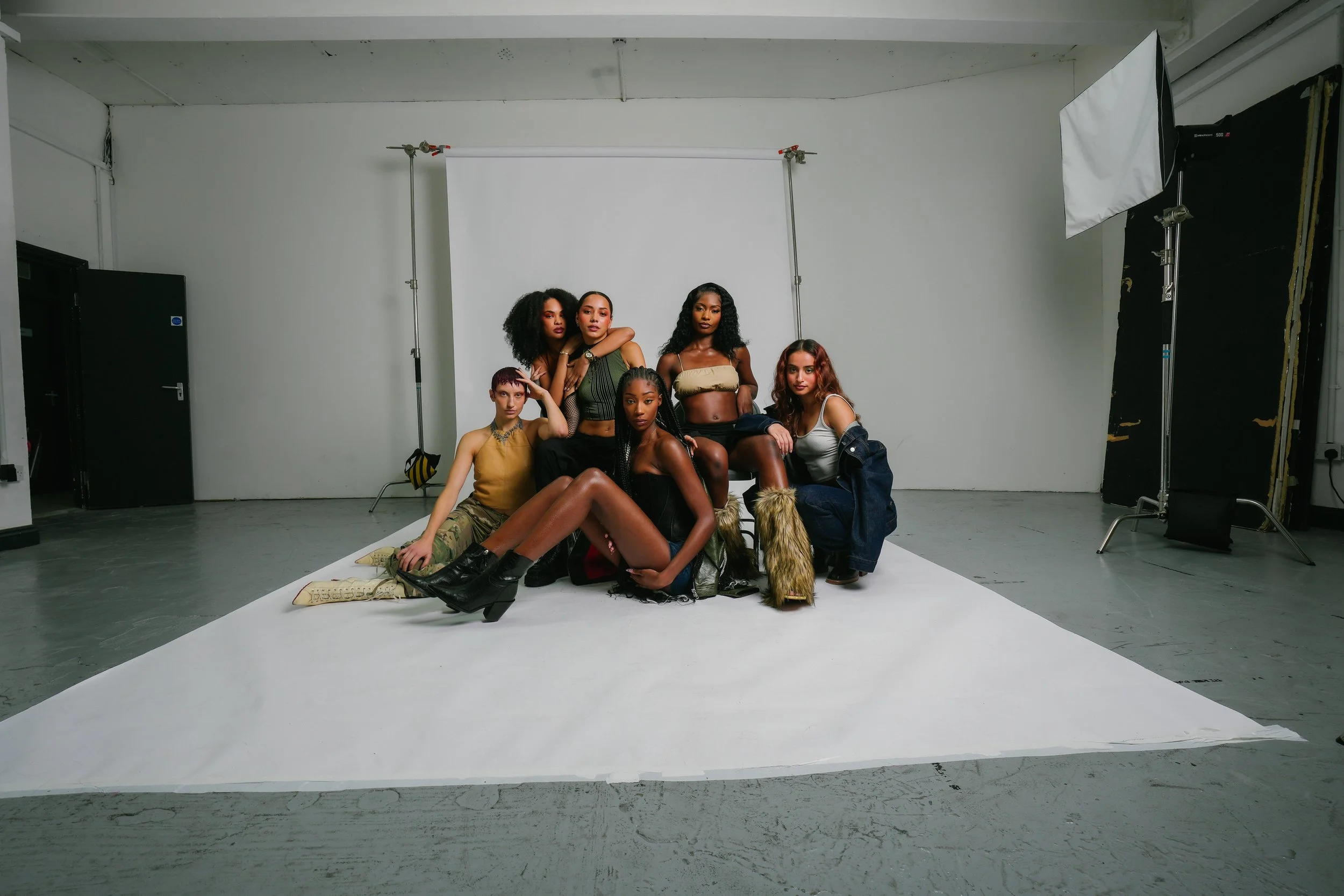 Six women posing together in a photography studio with a white backdrop. They are dressed in fashionable, diverse clothing and accessories, some sitting and others standing, all looking at the camera.