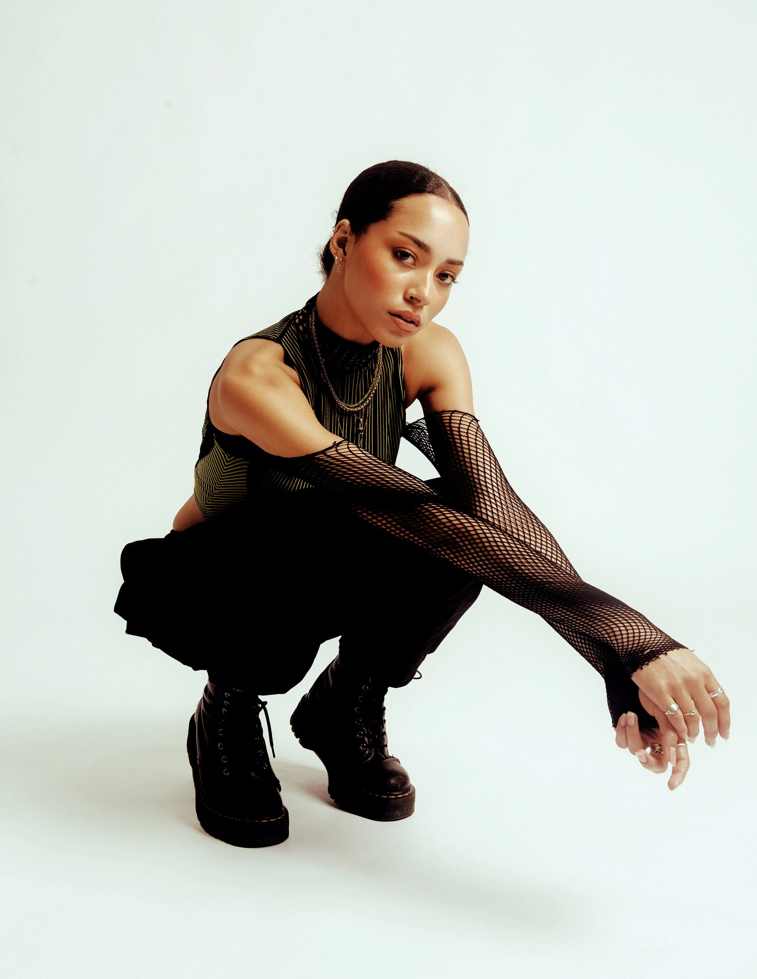 A young woman with short dark hair in a edgy outfit, including fishnet sleeves, black boots, layered necklaces, and rings, posing against a plain white background.