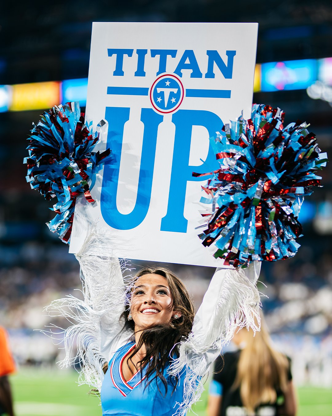 A Tennessee Titans cheerleader holding a sign that says 'TITAN UP' with pom-poms on either side during a football game.