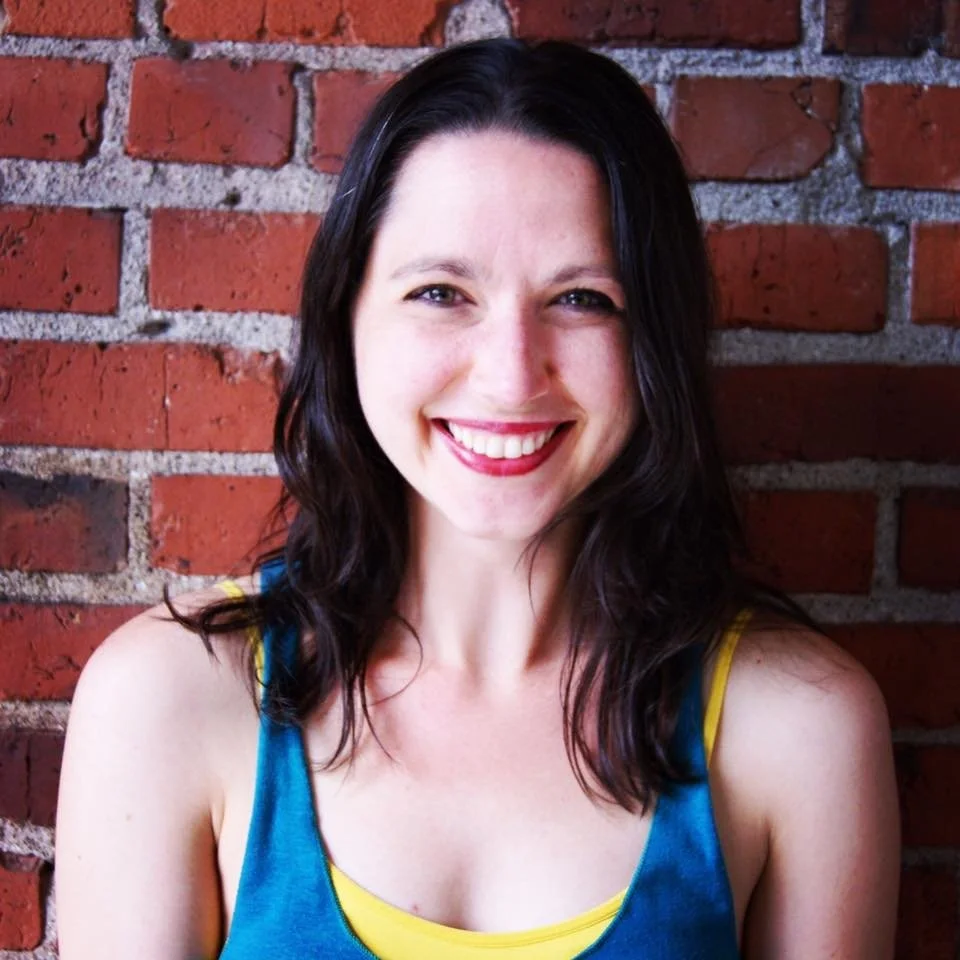 Smiling woman with dark hair wearing a blue tank top and yellow undergarment against a red brick wall.