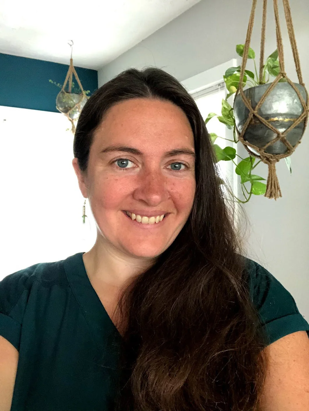A woman with long dark hair and blue eyes sitting indoors by a plant, wearing a black shirt, with a blue wall behind her.
