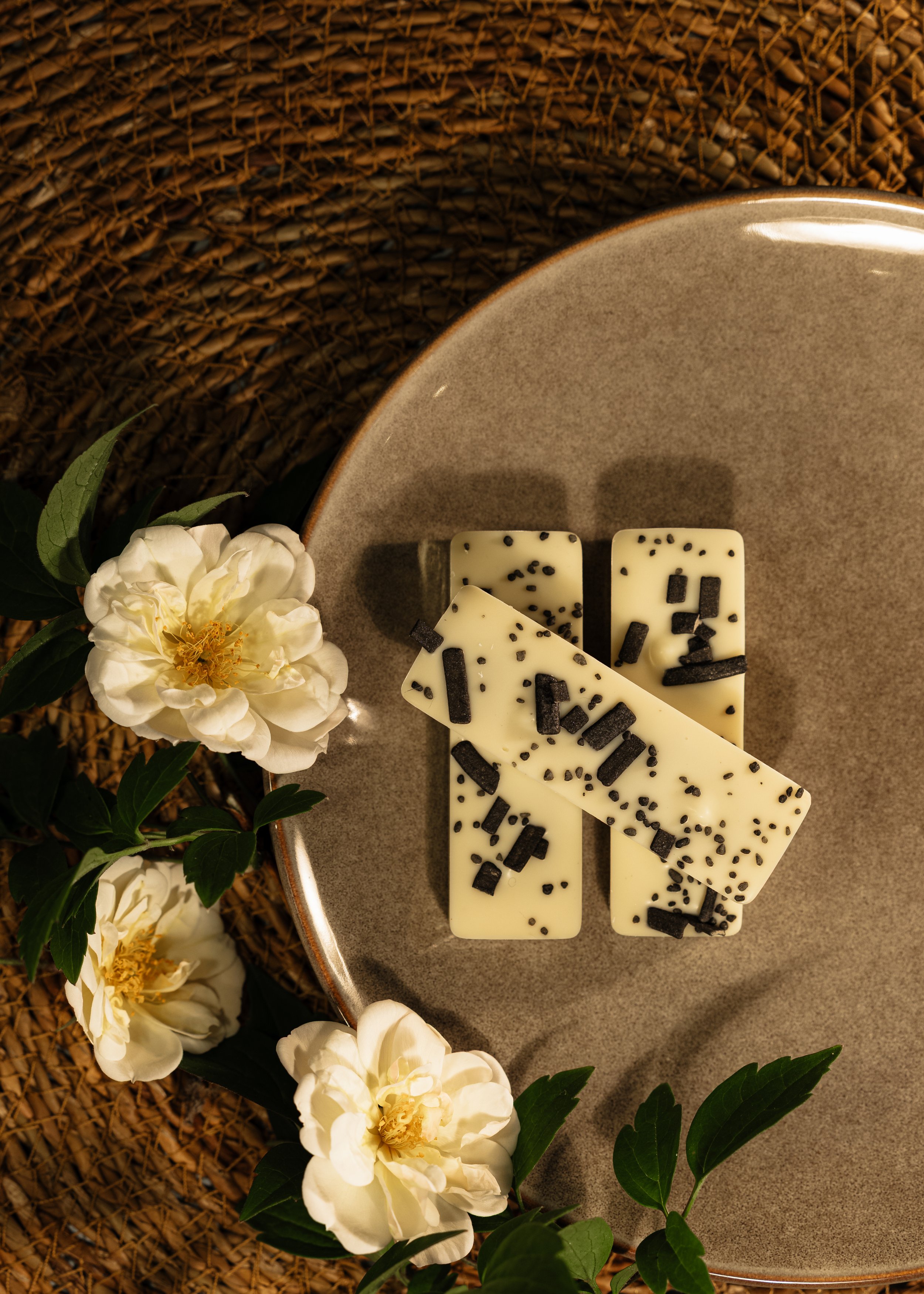 Hvide chokoladebarer med sorte sprøde stykker på en grå tallerken, omgivet af hvide blomster og grønne blade på en flettet barkunderlag.