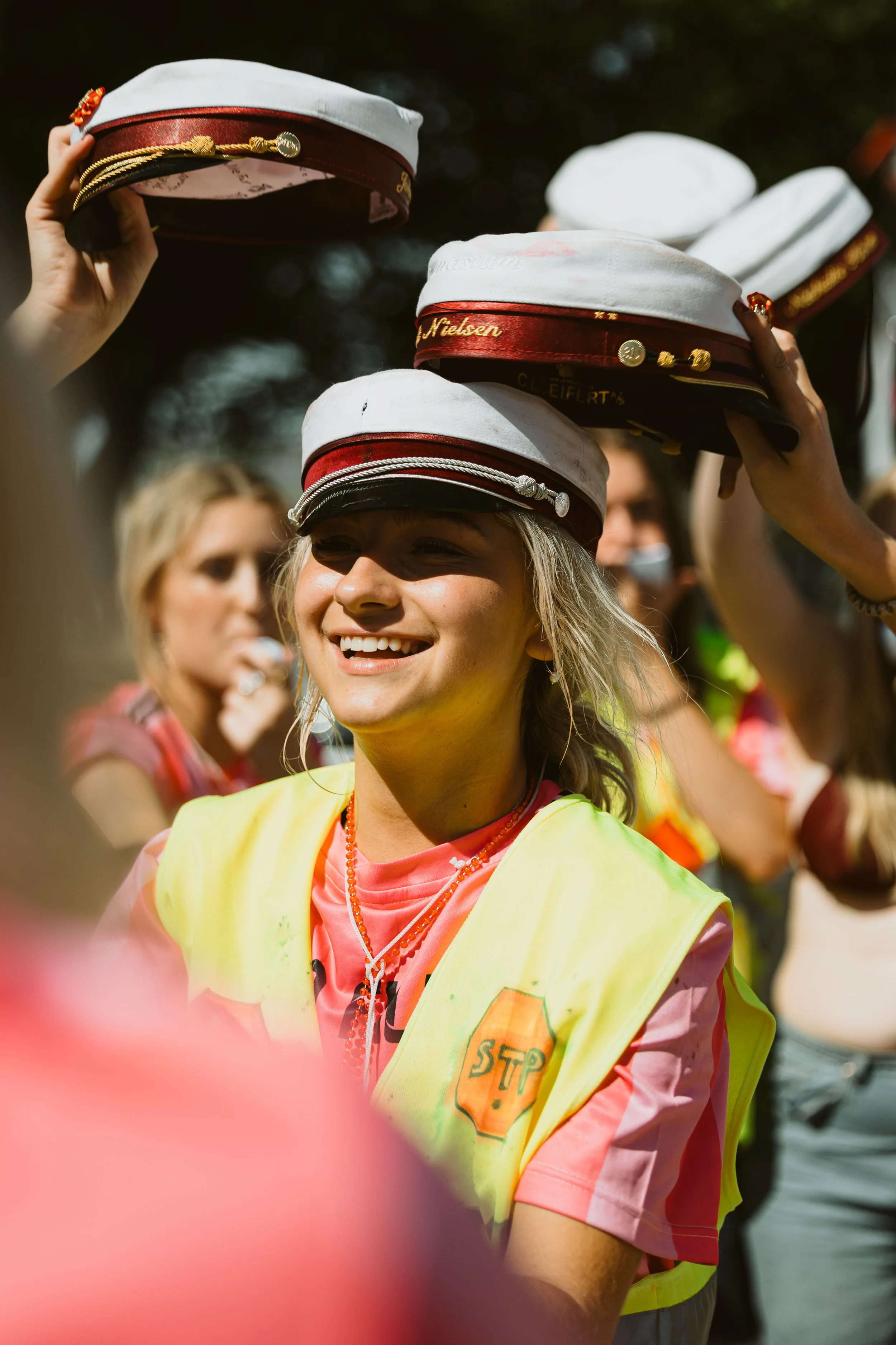 En gruppe unge mennesker, der holder huer over hovedet, tilsyneladende i feststemning uden for, med fokus på en smilende kvinde iført en gule vest og en rosa t-shirt.