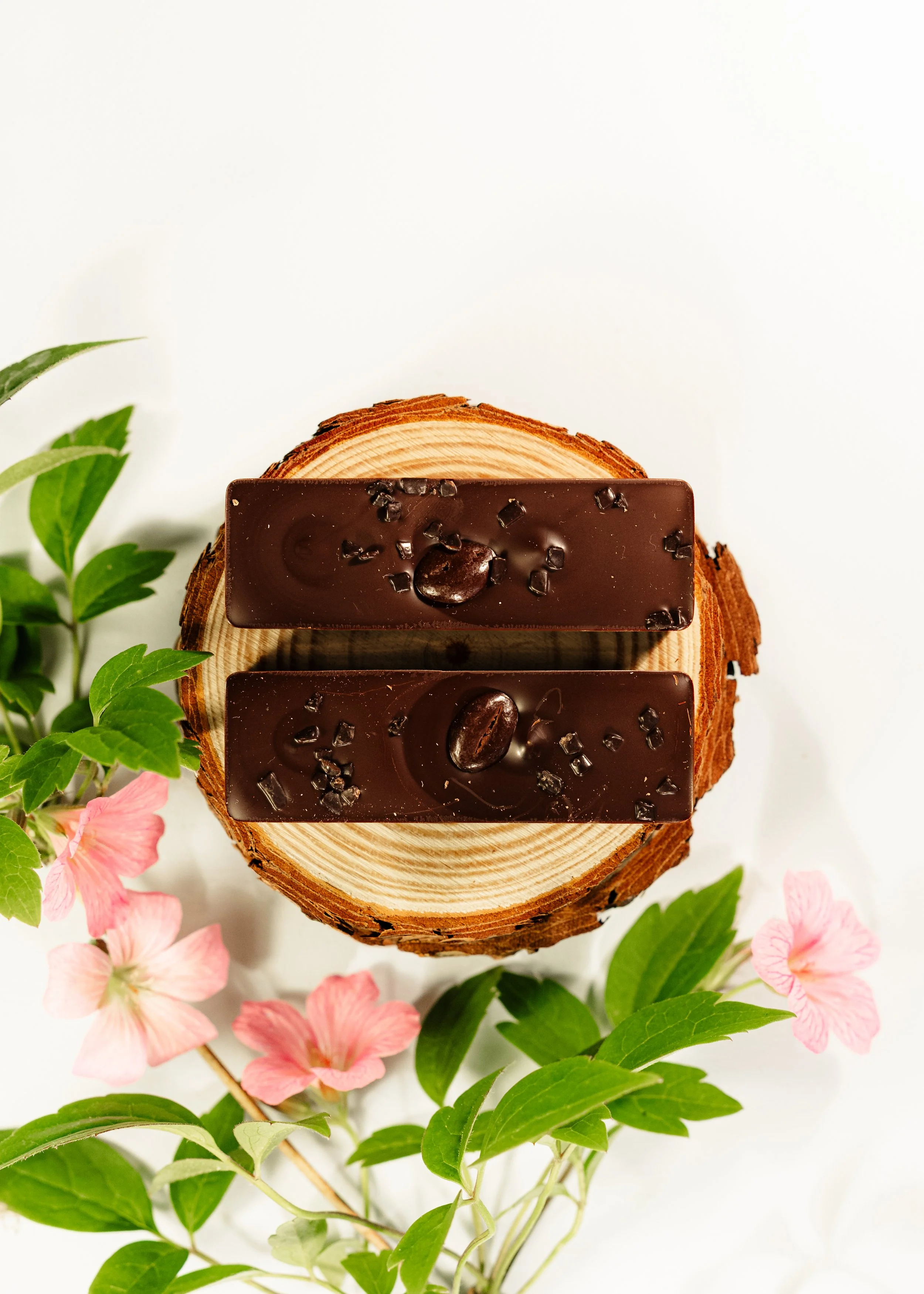 Nærbillede af to chokoladeplader med kokos- og kaffebønner, placeret på et skive af træ, omgivet af grønne blade og pink blomster.