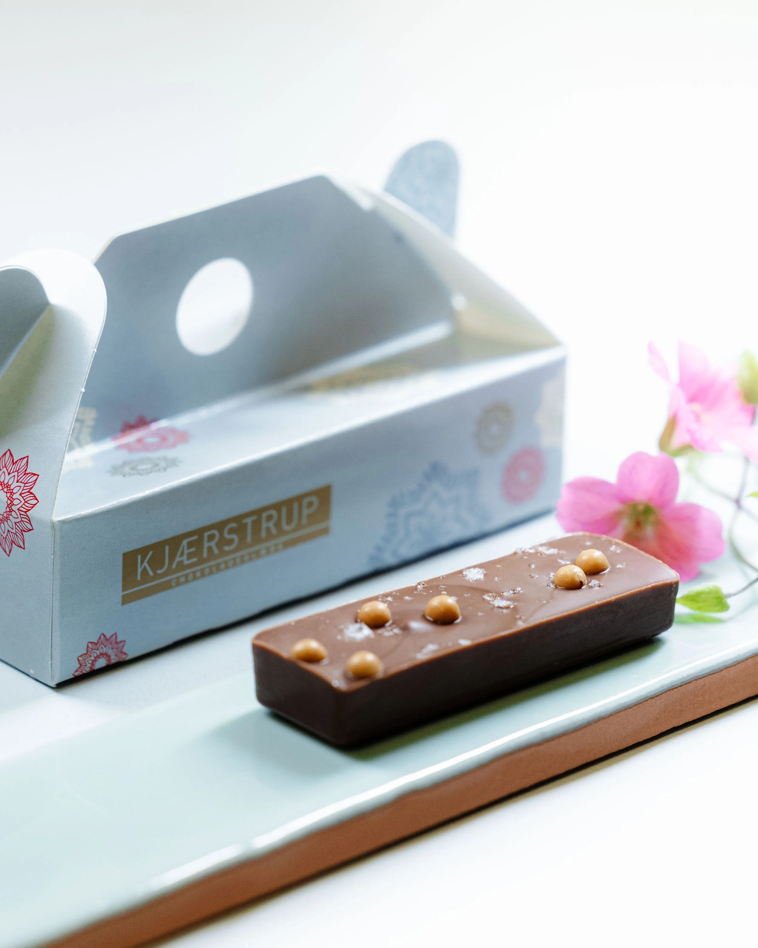 En chokoladebar med nødder på en hvid tallerken, ved siden af en pakke med kys og nogle pink blomster.
