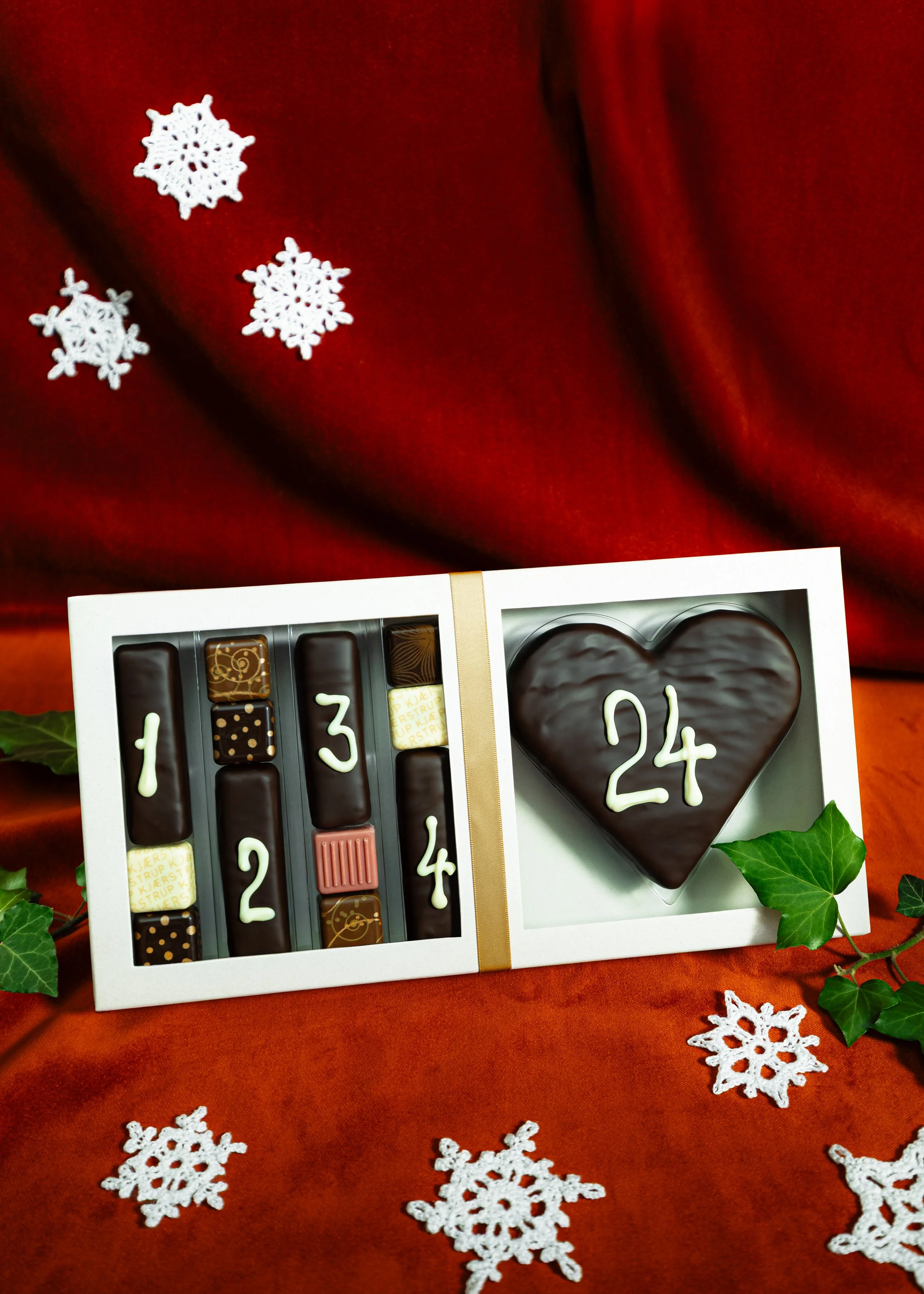 En gaveæske med chokolade, der er pyntet med hvide julemotiver, og en lysende hjertefigur med nummer 24, omgivet af snefnug og grønne blade på en rød dug.