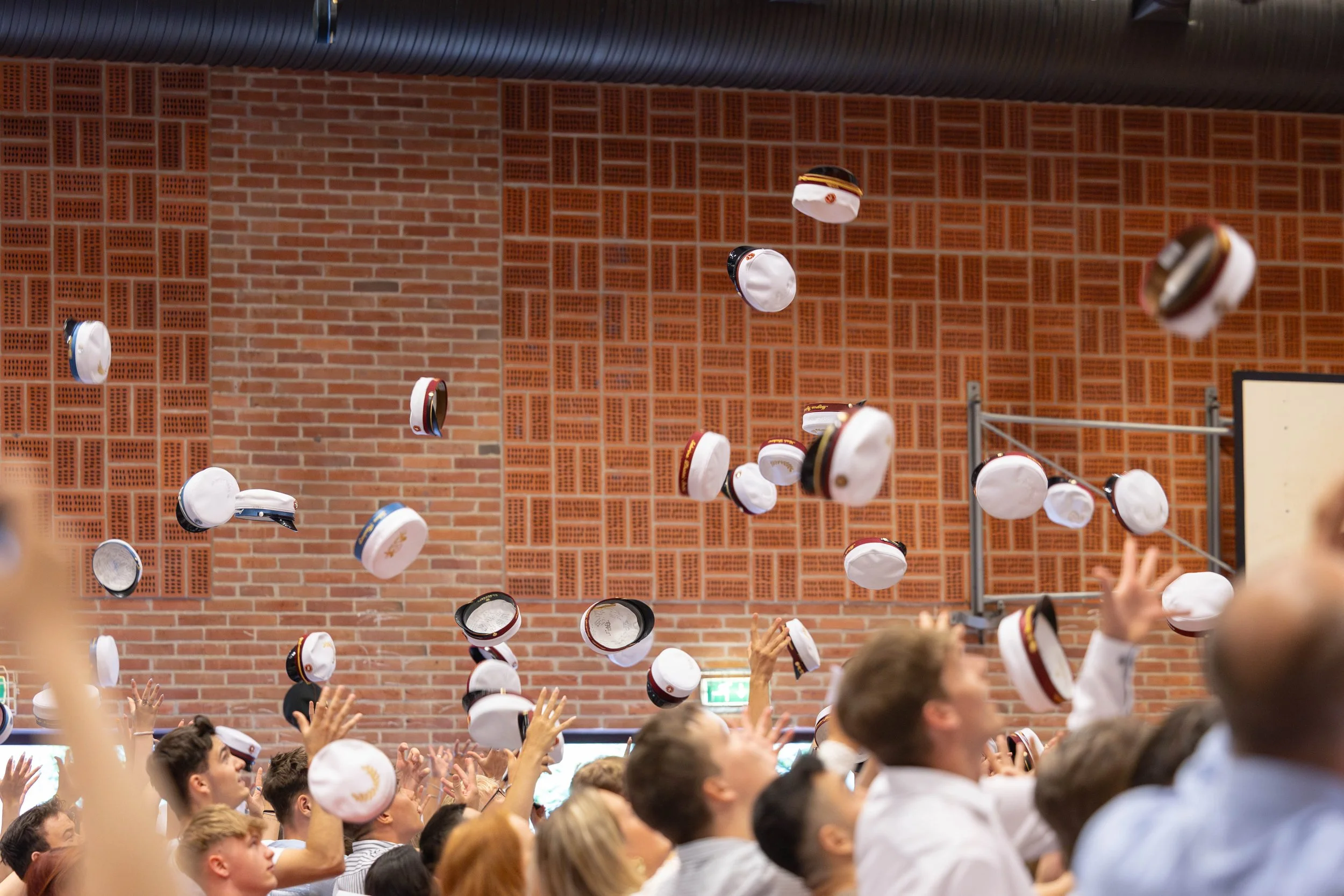 Folk i graduationhatte kaster hatten op i luften indendørs med murstensvæg i baggrunden.