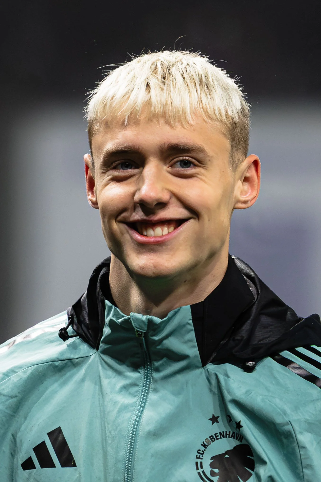 En ung mand smiler bredt med blond hår og blå øjne, iført en lysegrøn Adidas jakke med sorte og hvide detaljer, og en sort træningsjakke med en logo, sandsynligvis på en sportsbegivenhed.