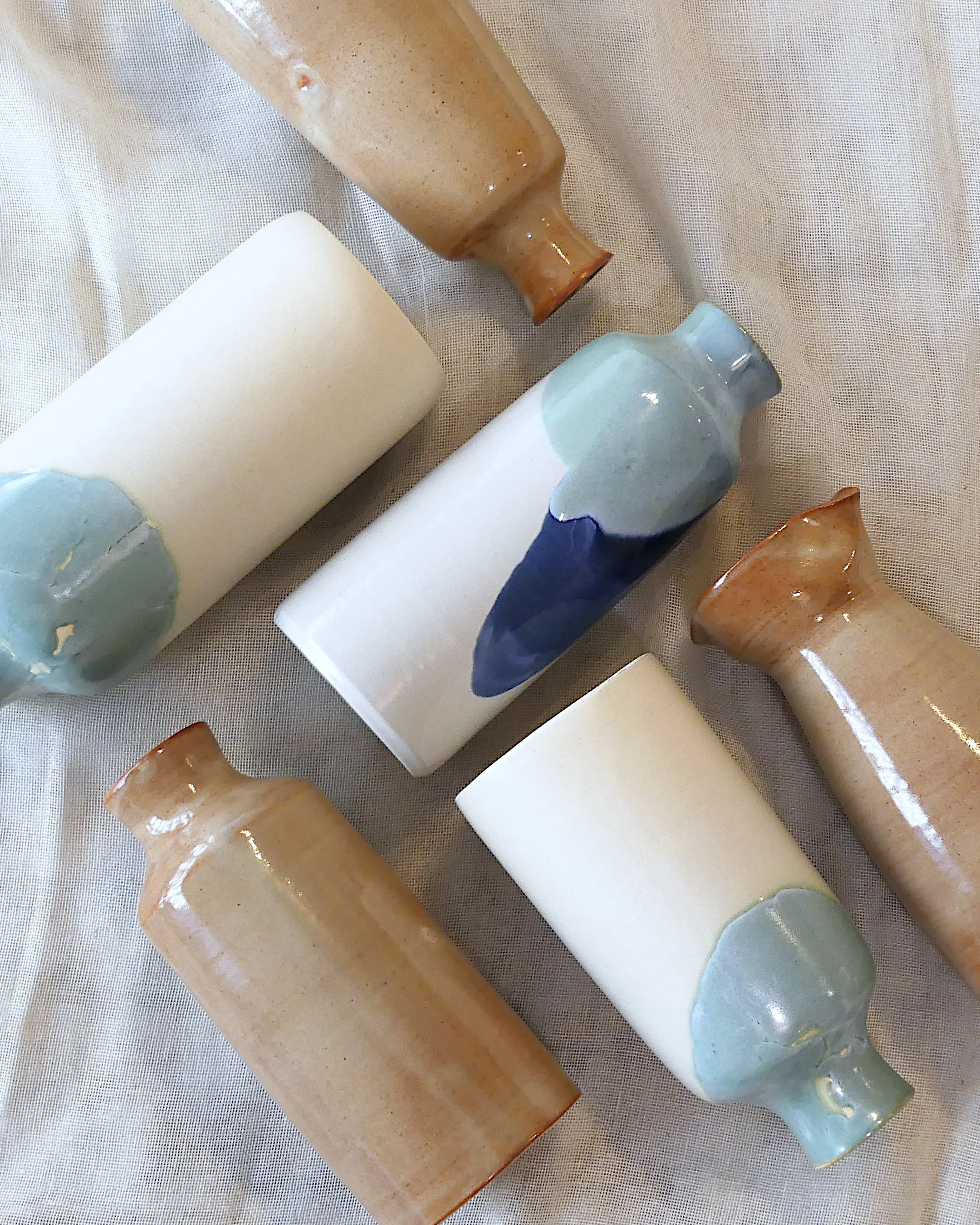 6 bouteilles en céramiques vue de dessus, posées sur une nappe en toile écrue. Les bouteilles sont blanches, sables, bleues, vertes et ambrées.