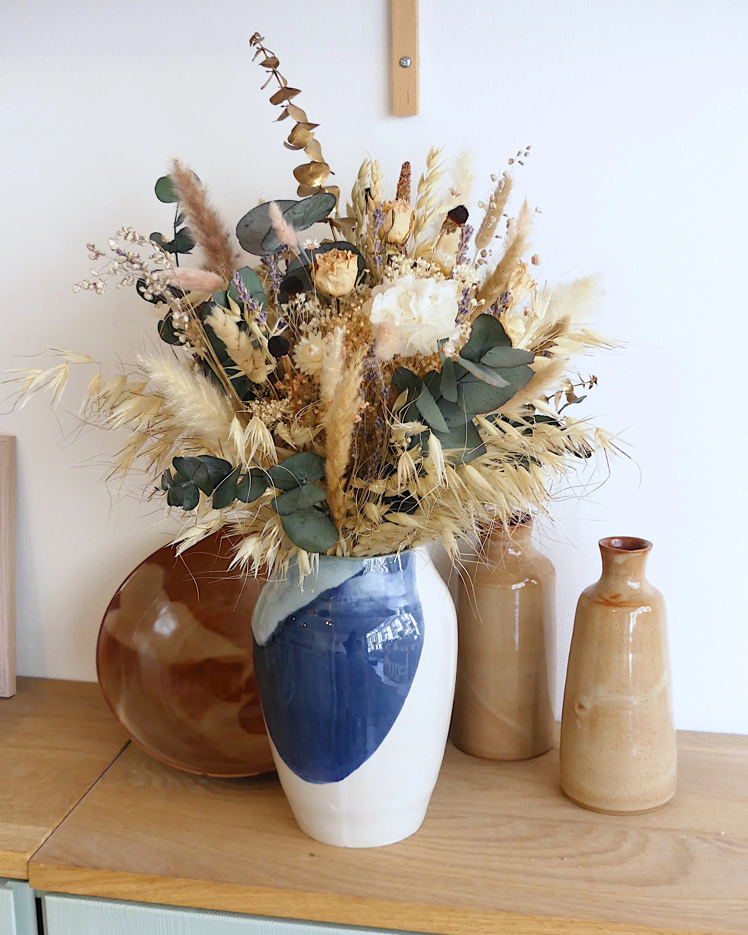 Au centre, un grand vase blanc bleu et vert contenant un bouquet de fleurs séchées. Autour des bouteilles en céramique de couleurs ambrées et un saladier dans les même tonalités de couleur.