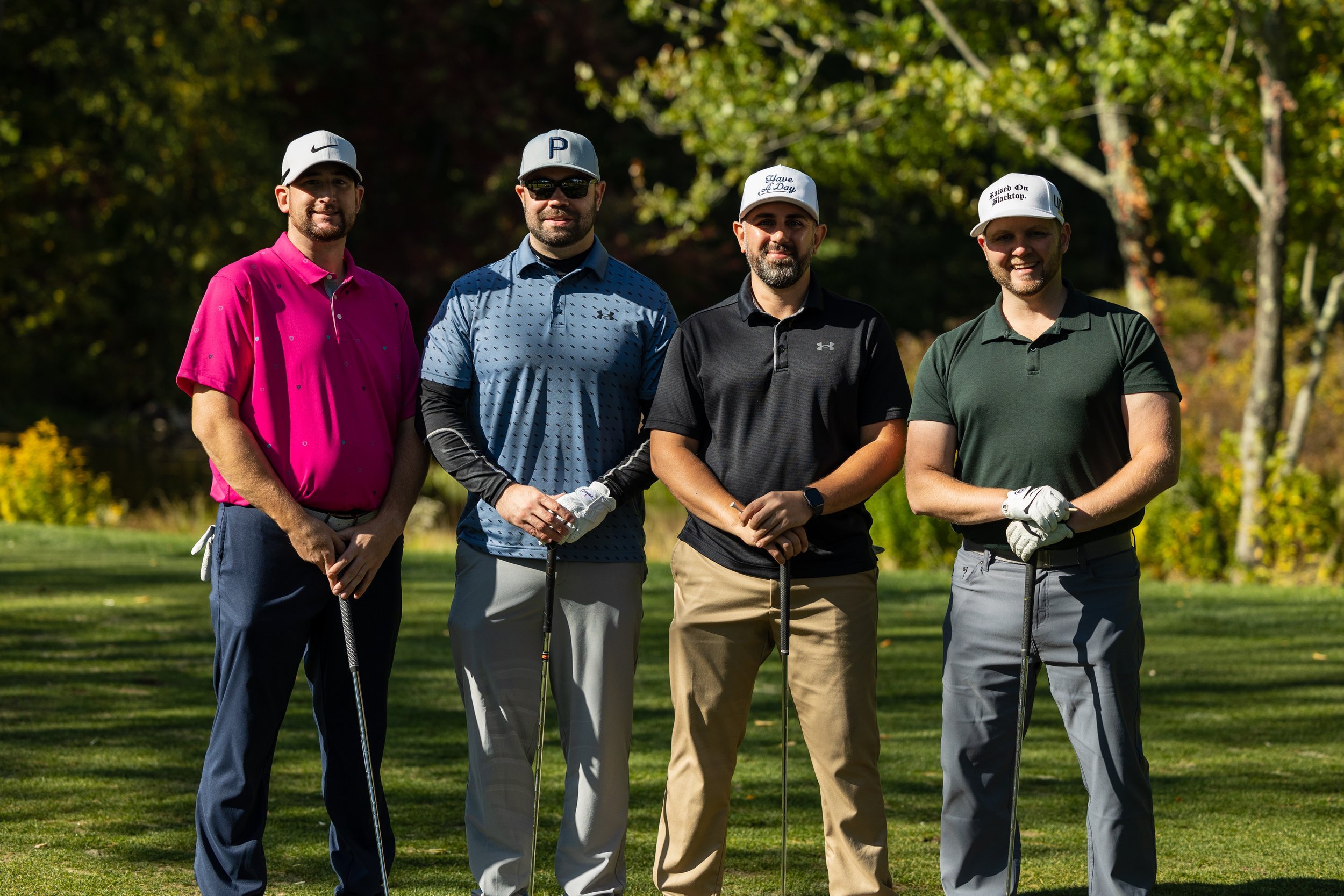 Four men standing on a golf course holding golf clubs, wearing casual golf attire and hats, with trees in the background.