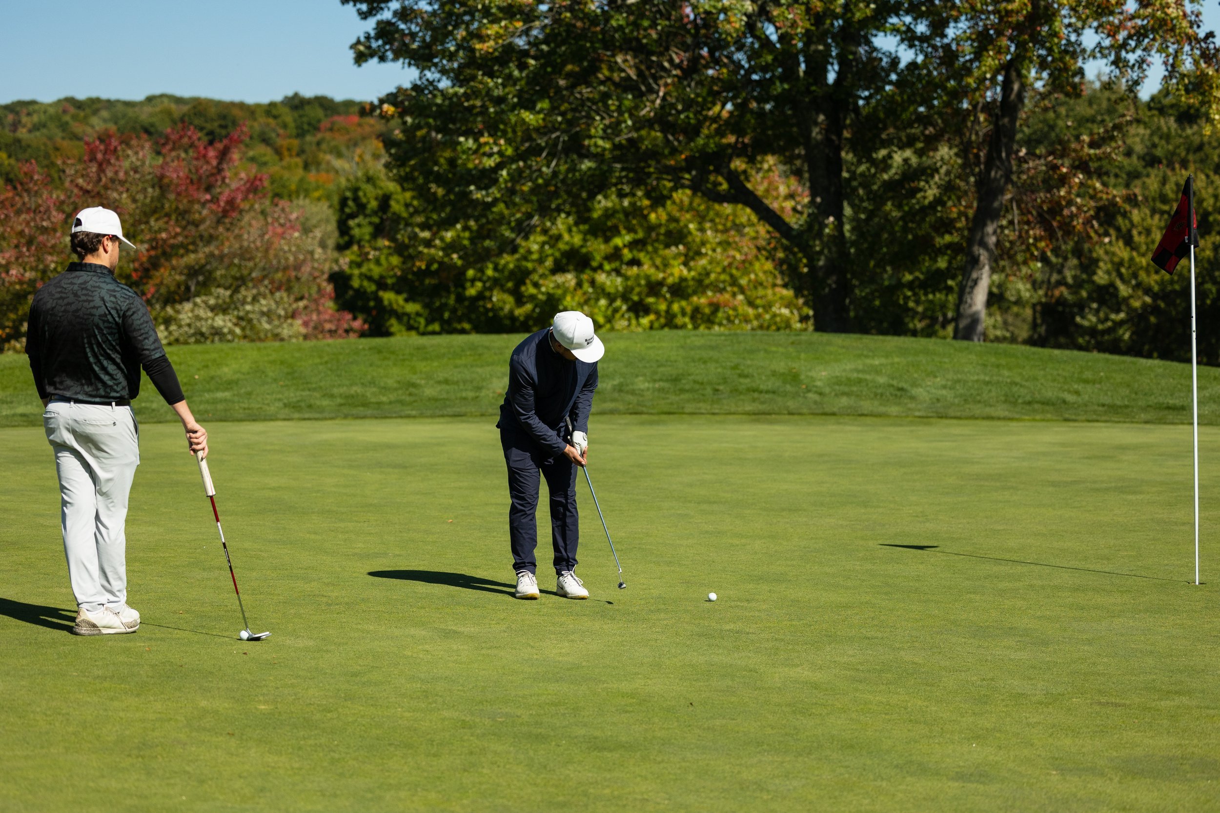 Two golfers on a green, one putting, with golf flag nearby and trees in the background.