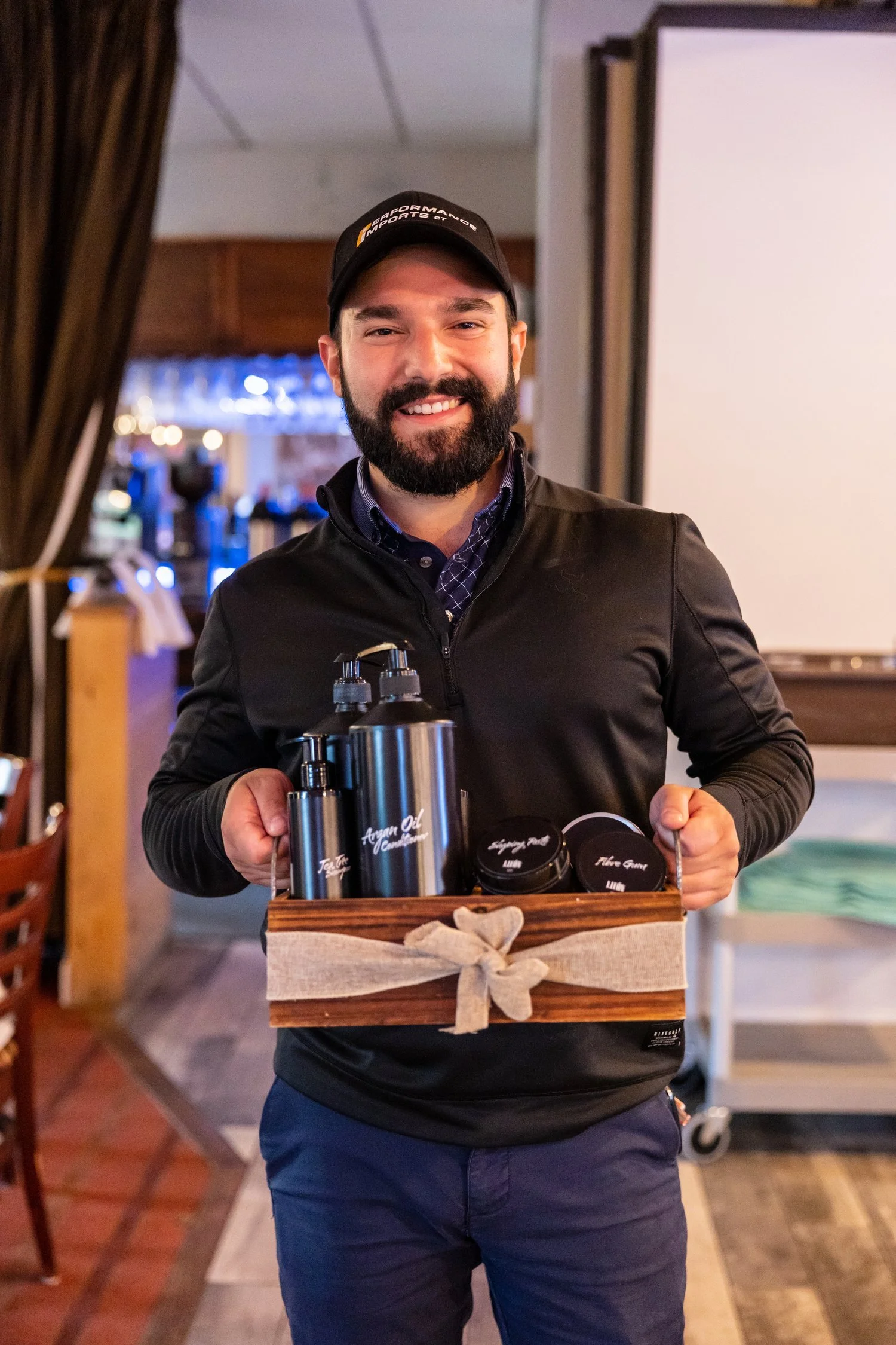 Man holding a gift set with hair and skincare products, wearing a black cap and jacket, smiling.