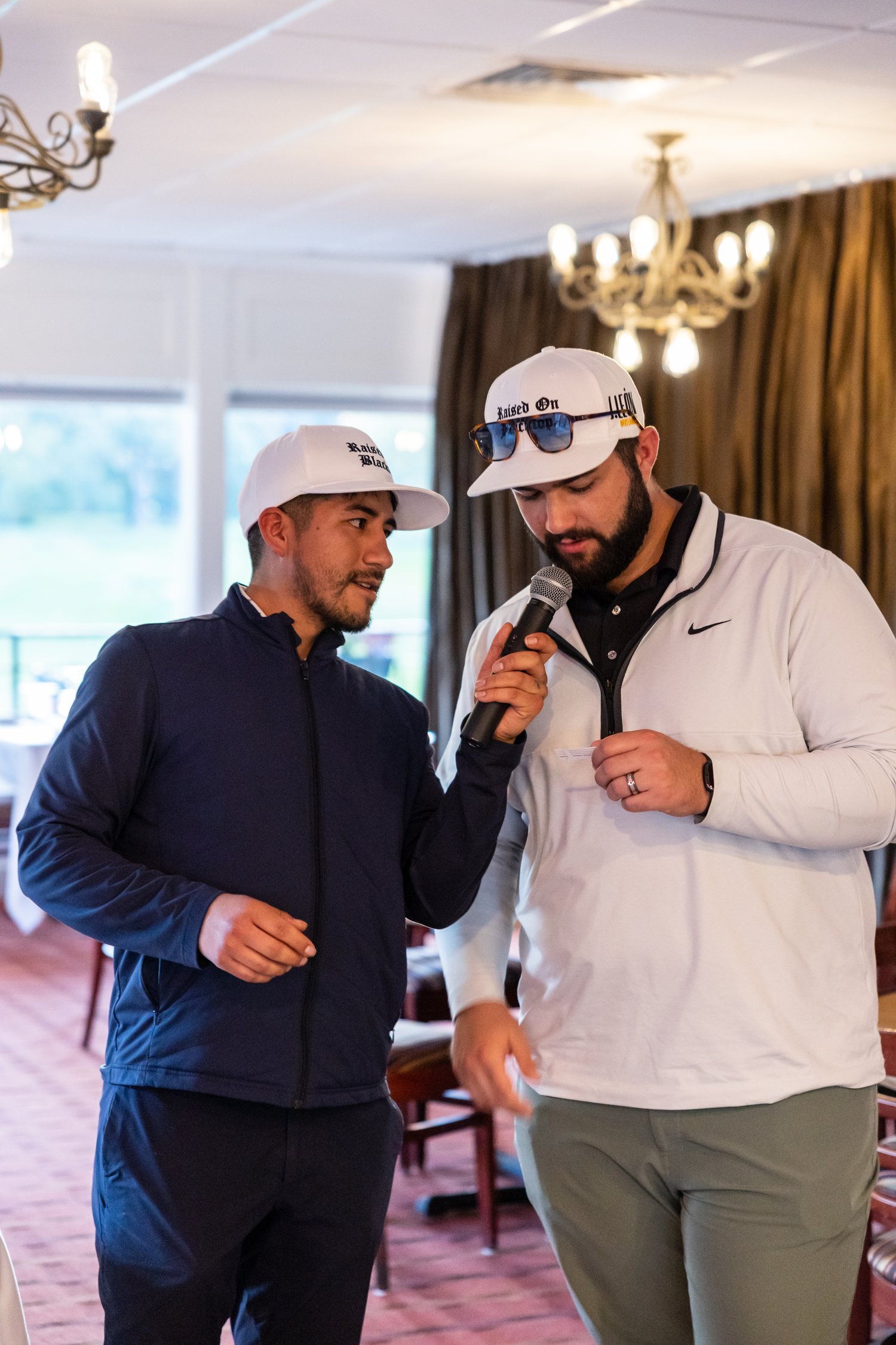 Two men wearing white caps are indoors, one holding a microphone and speaking while the other listens. They are in a room with chandeliers and brown drapes.