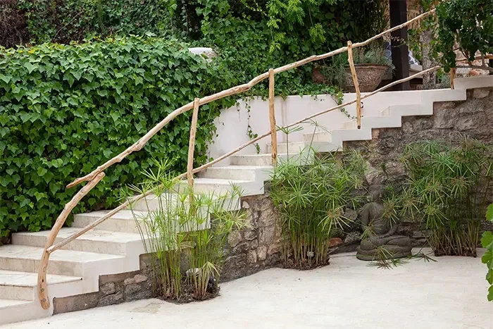Outdoor stone staircase with wooden handrail, surrounded by lush greenery and potted plants.