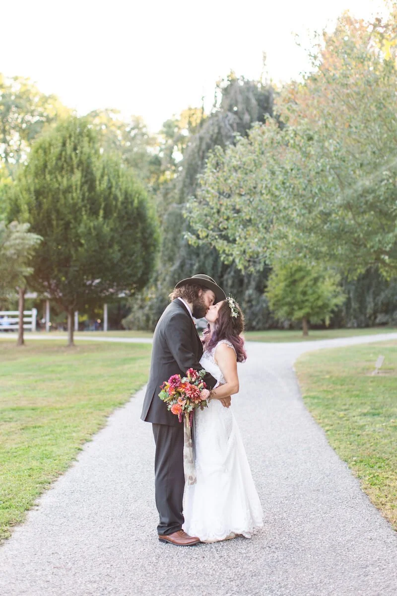 The bride with a crown of flowes and her bouqet kissing the groom on a path with trees behind them in the distance.jpg