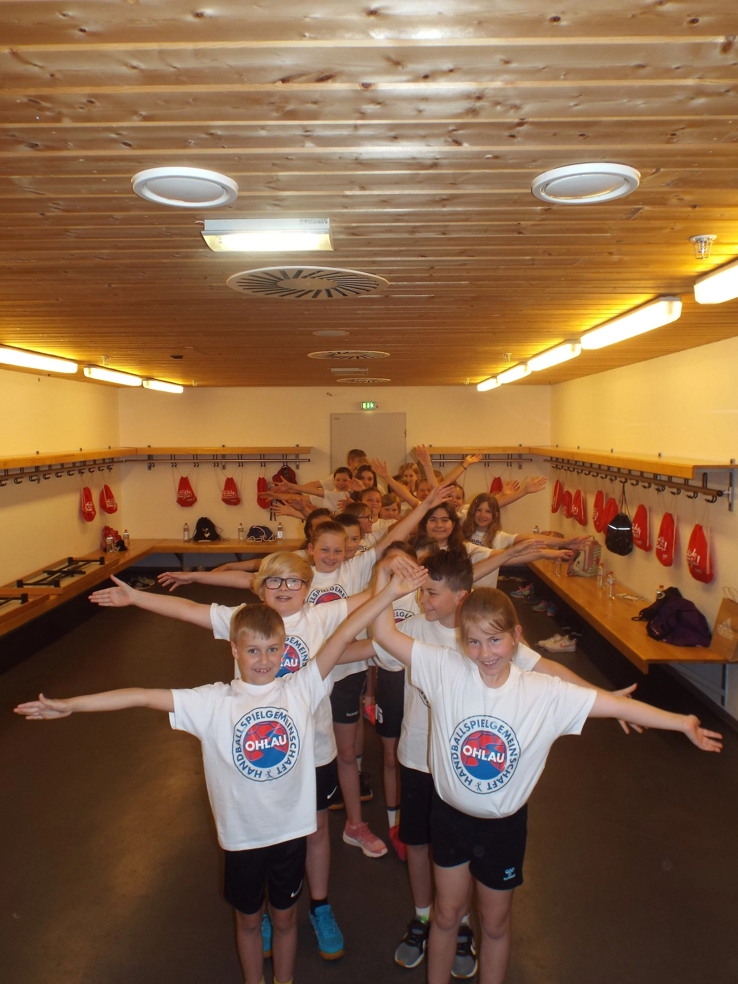 Kinder in einer Umkleidekabine mit weißen T-Shirts, die auf den Schultern "OHL“ stehen haben. Sie posieren in einer Reihe mit ausgestreckten Armen. Im Hintergrund sind rote Taschen an der Wand.