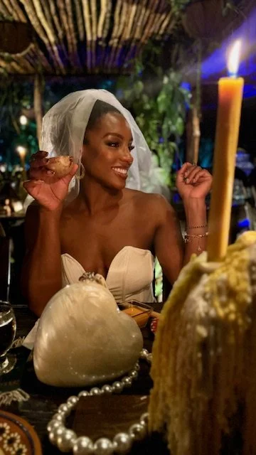 A woman dressed in white with a veil, smiling at a dinner table, holding a piece of bread, in a tropical or outdoor setting with candlelight and greenery.