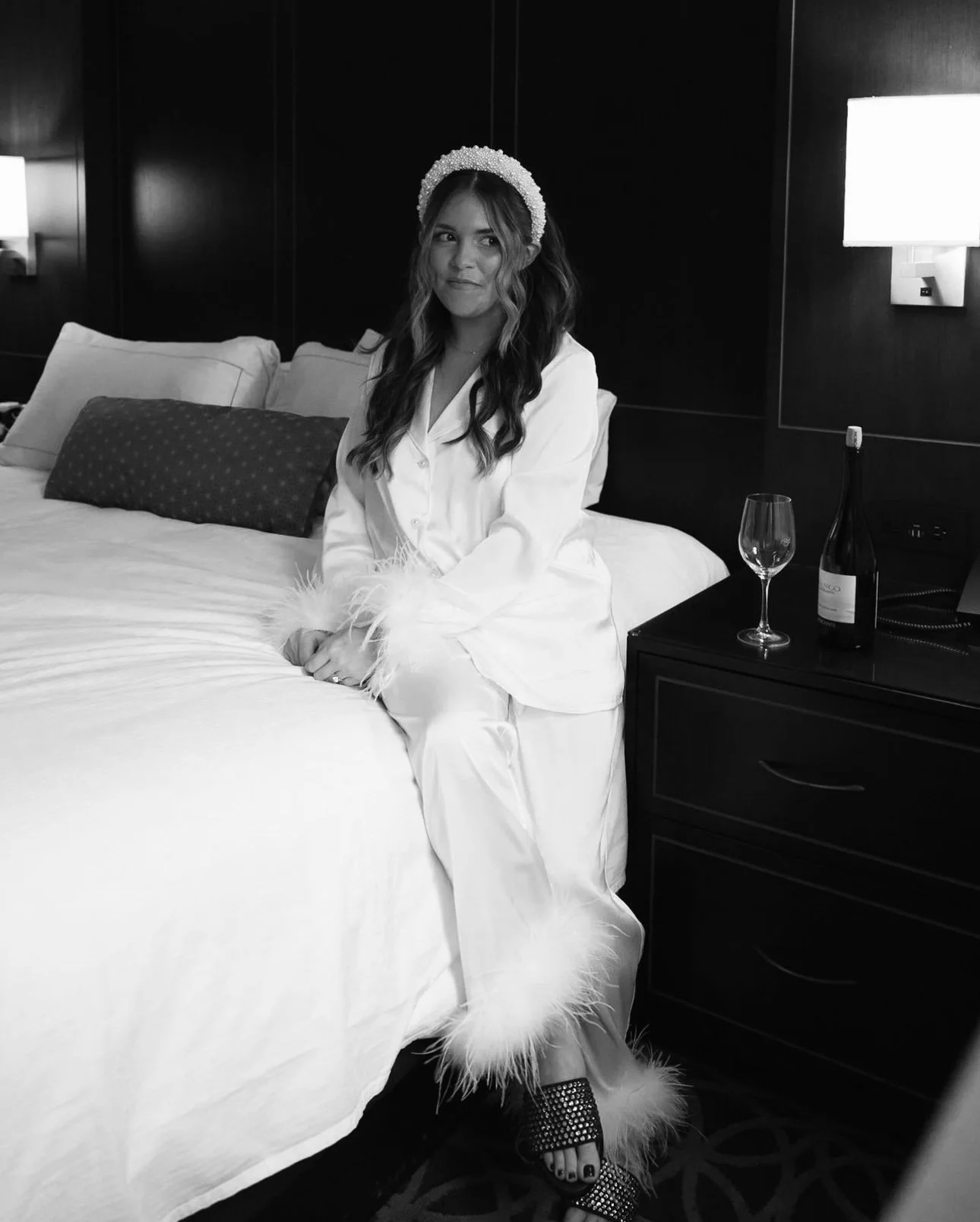 A woman sitting on the edge of a bed in a hotel room, wearing satin pajamas with feather trim, a pearl headband, and high-heeled sandals, with a champagne bottle and glass on a bedside table.