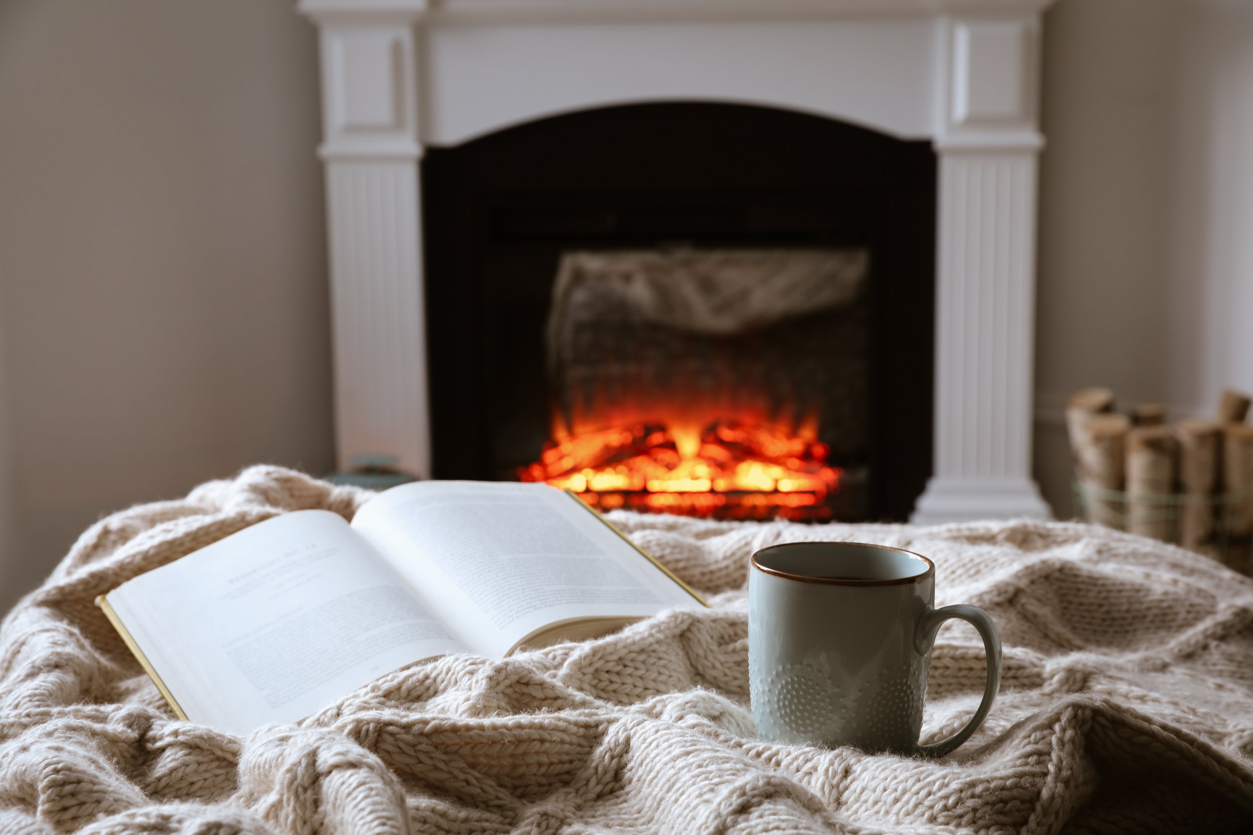 A cozy scene with an open book, a ceramic mug, and a knitted blanket on a bed in front of a fireplace with glowing embers