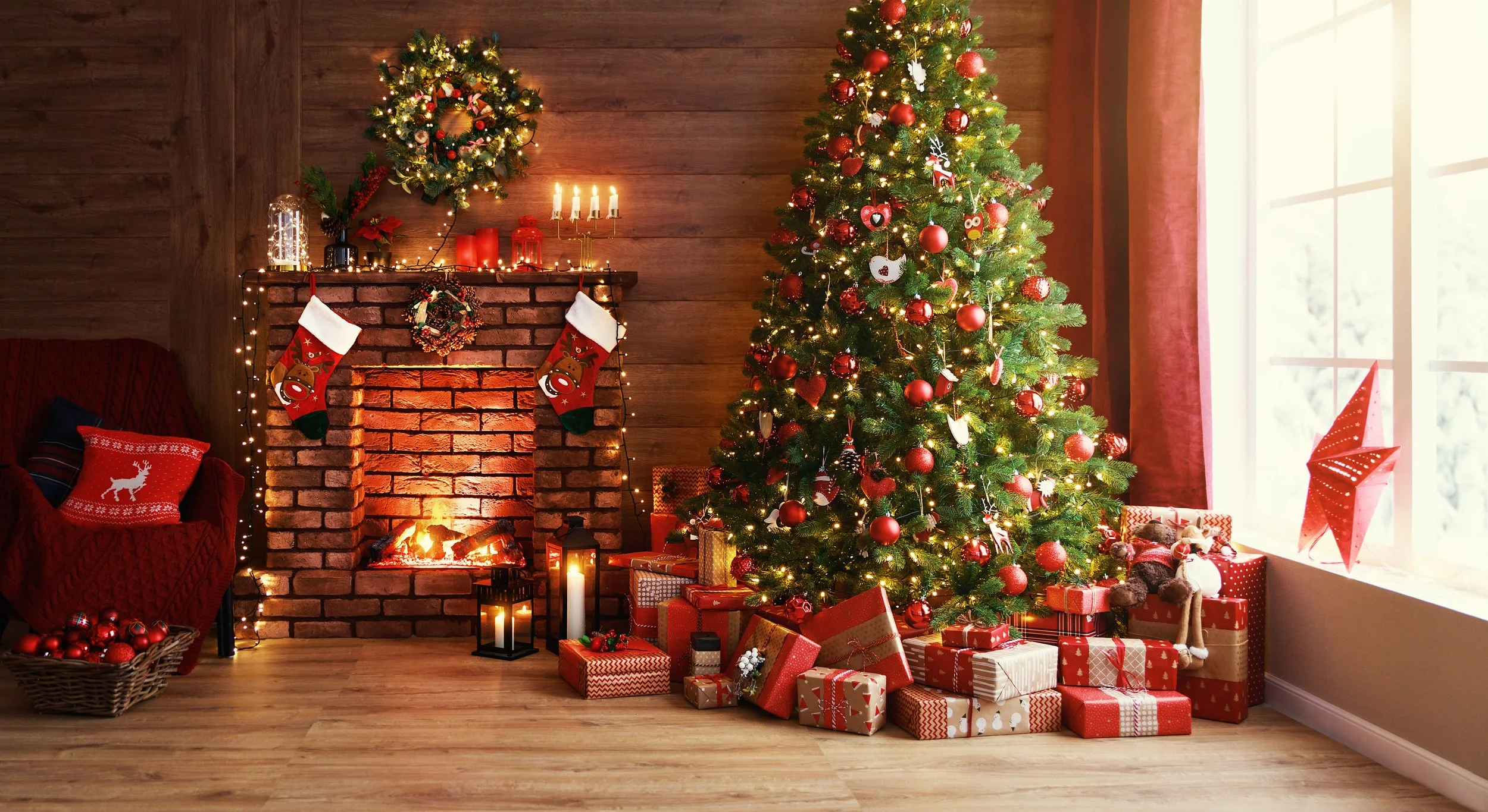 Christmas tree decorated with red ornaments and lights, surrounded by wrapped presents, placed in a cozy living room with a brick fireplace, stockings hanging, and festive decorations including a wreath, candles, and a paper star on the window.