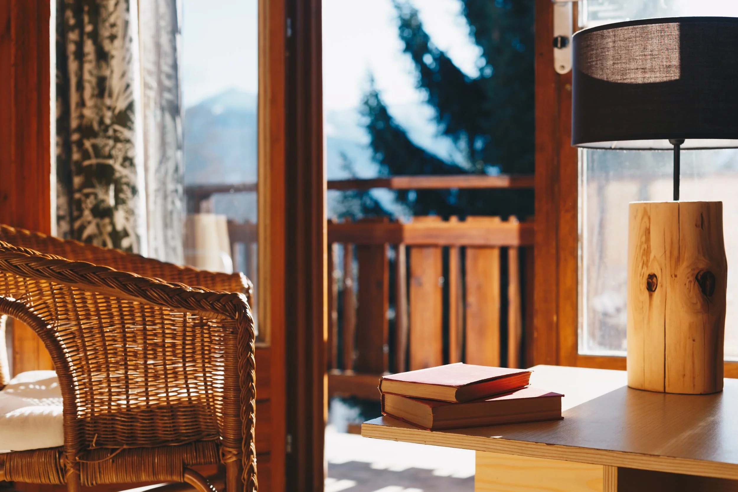 A cozy indoor space with a wicker chair, a wooden table with three books, a table lamp with a wooden base and black lampshade, and large windows showing an outdoor deck and snow-covered trees in the background.