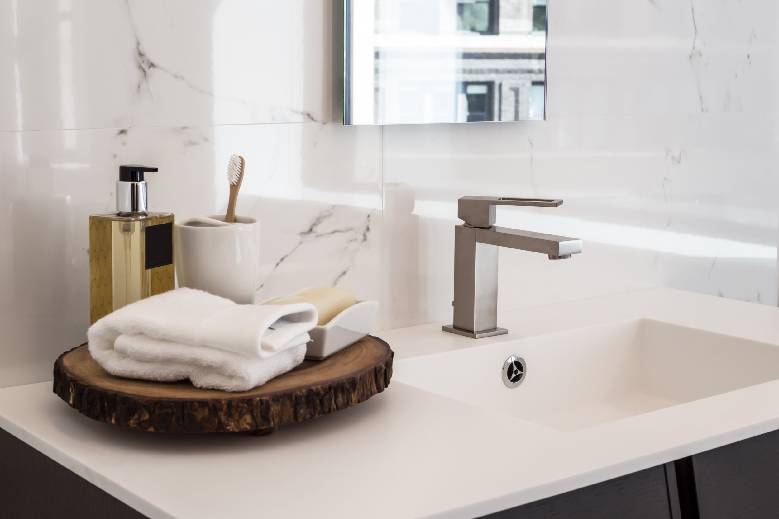 Bathroom sink area with a modern silver faucet, a wooden tray holding white towels, a soap dispenser, a toothbrush in a cup, and a mirror above.