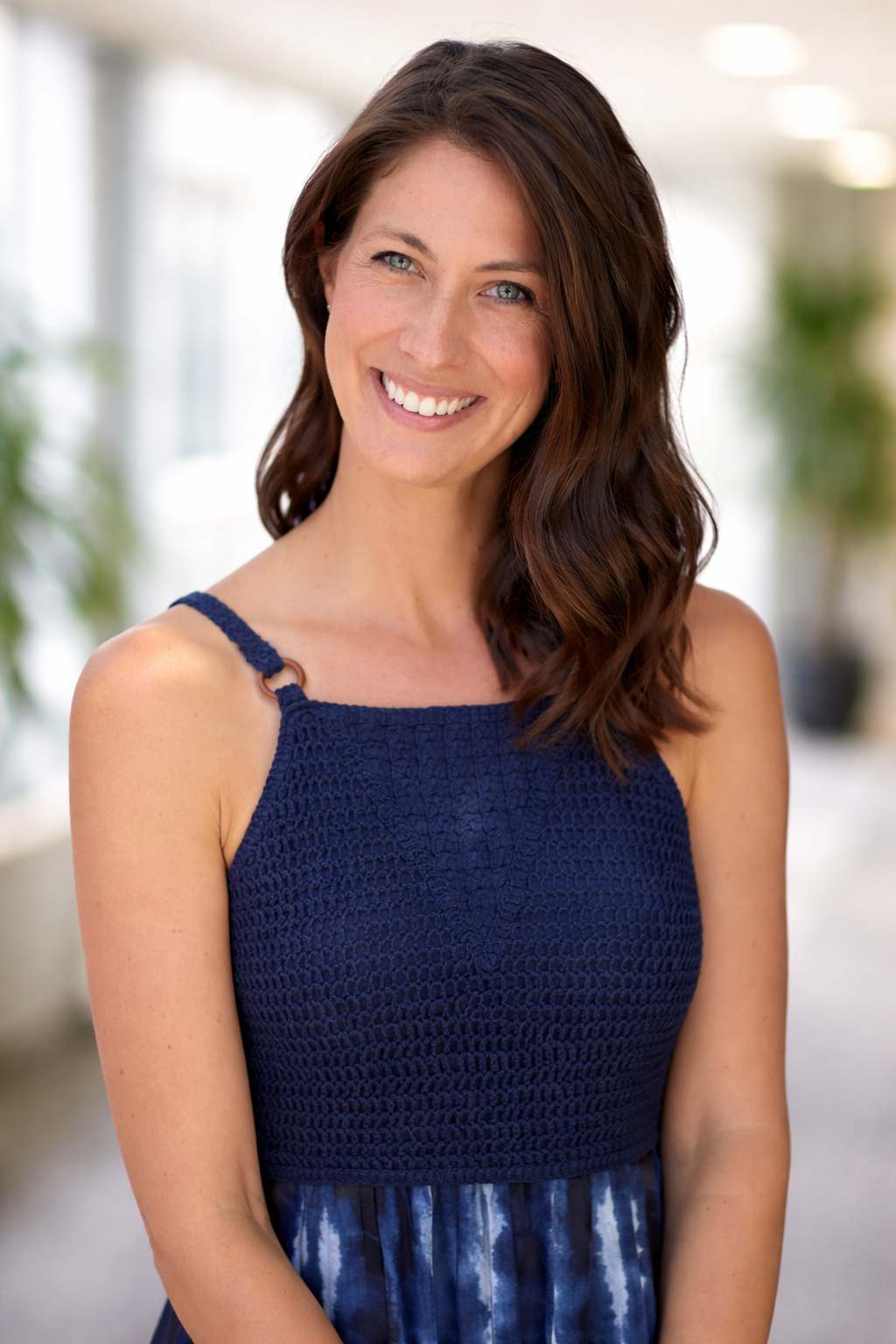 A woman with shoulder-length wavy brown hair, blue eyes, and light skin, smiling in focus against a blurred background with greenery.