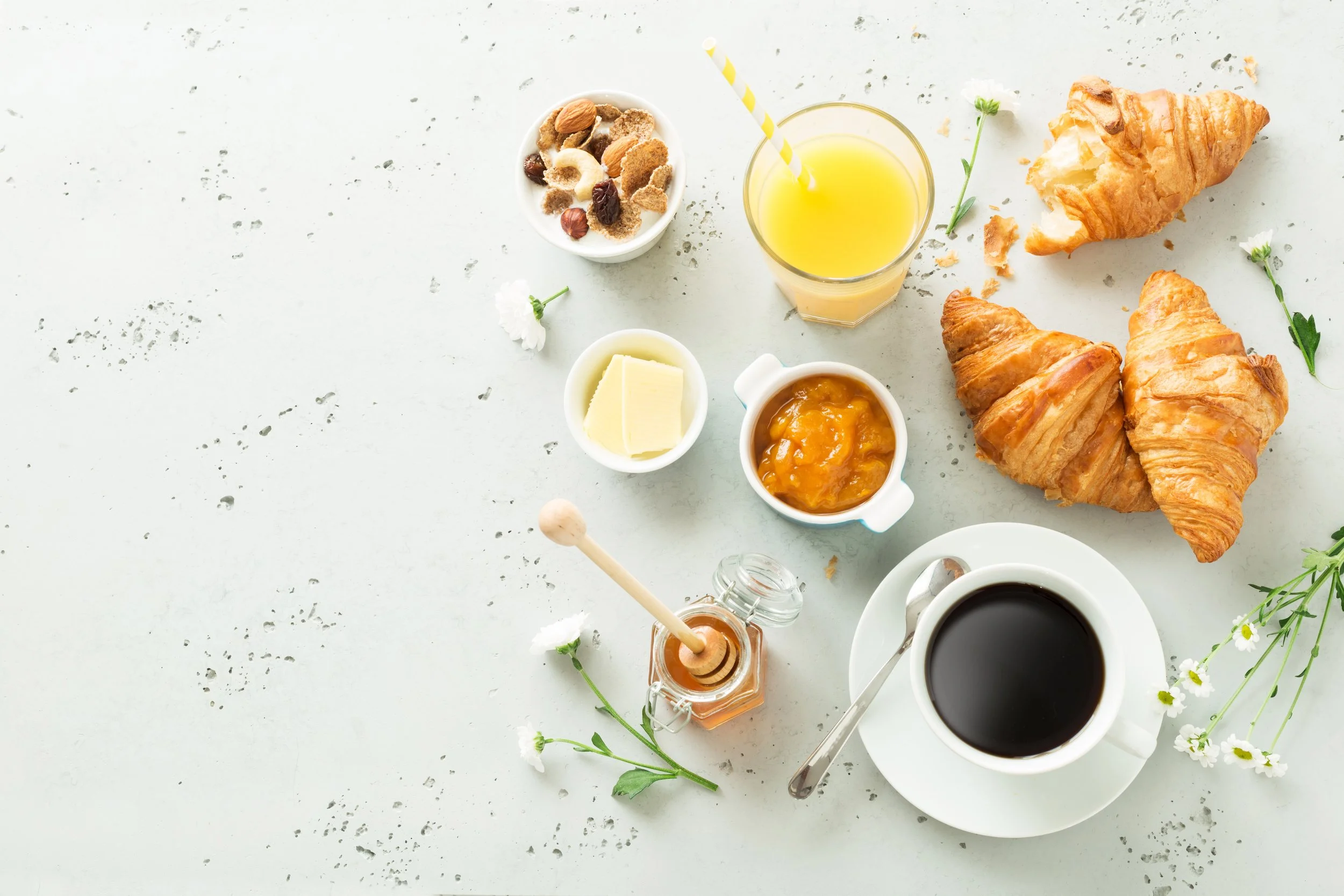 Breakfast setup with croissants, coffee, orange juice, honey, butter, jam, cereal, and flowers on a white surface.