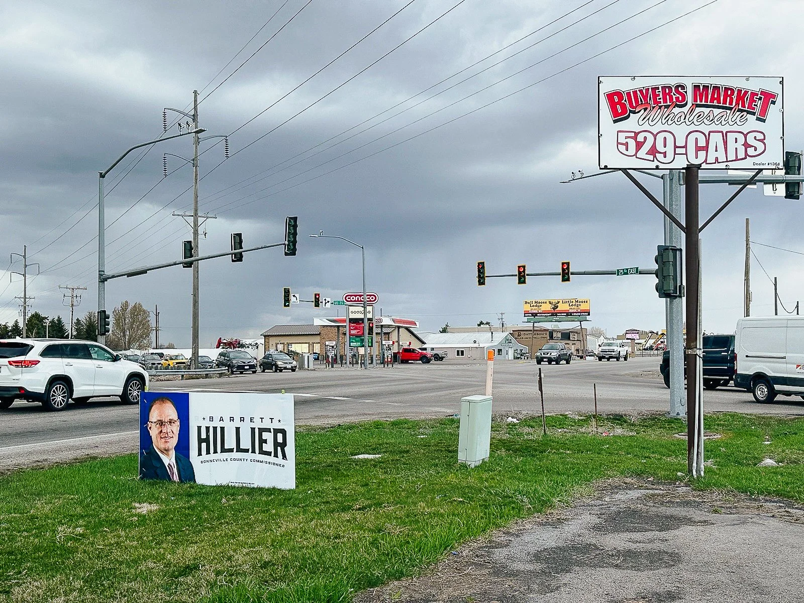View of a busy intersection with a large billboard advertising buyers market wholesale cars and a political campaign sign for Barrett Hillier, all under a cloudy sky.