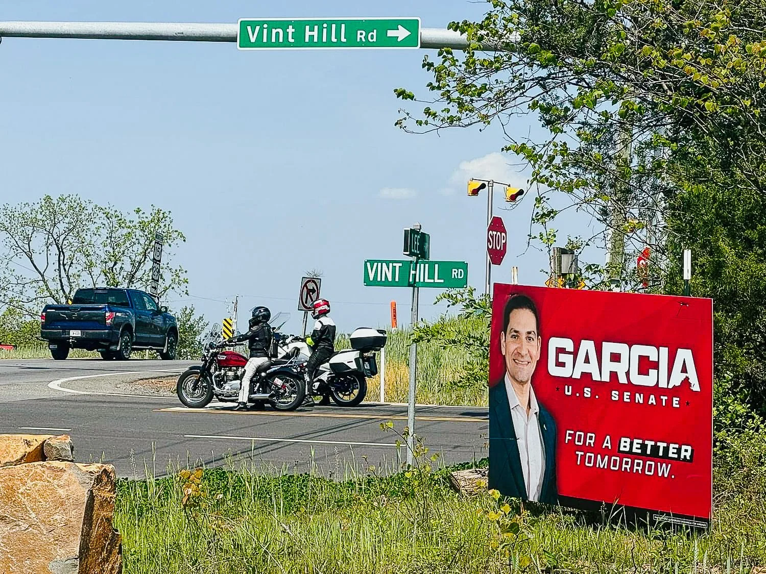 A red political campaign sign for Garcia, U.S. Senate, featuring a smiling man in a suit, with the slogan 'For a Better Tomorrow,' placed beside a highway with motorcycles and a car. The scene includes street signs for Vint Hill Road and a stop sign,