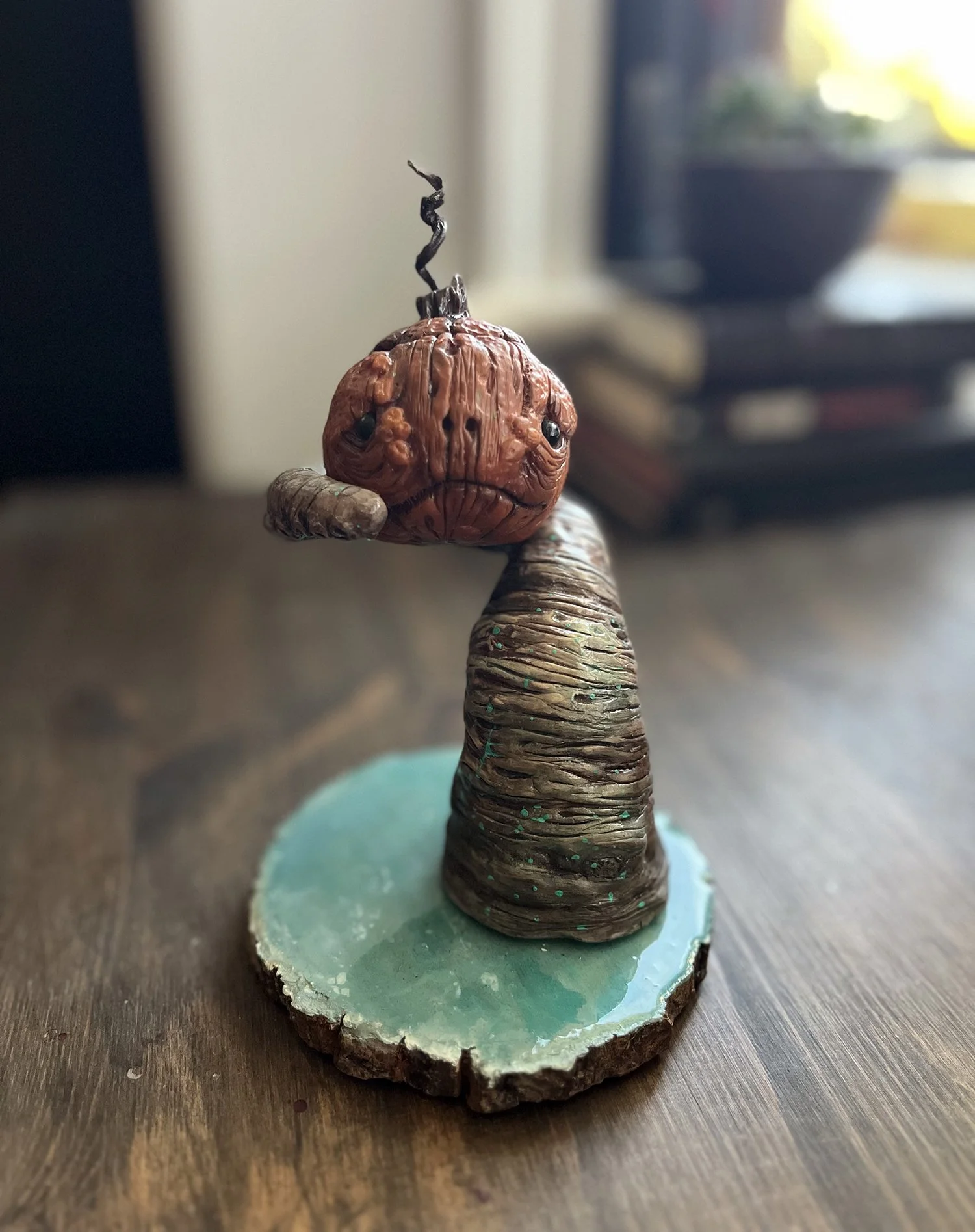 A small sculpture of a winding tree with a pumpkin head on a wooden base, with a blurry background of books and a potted plant.