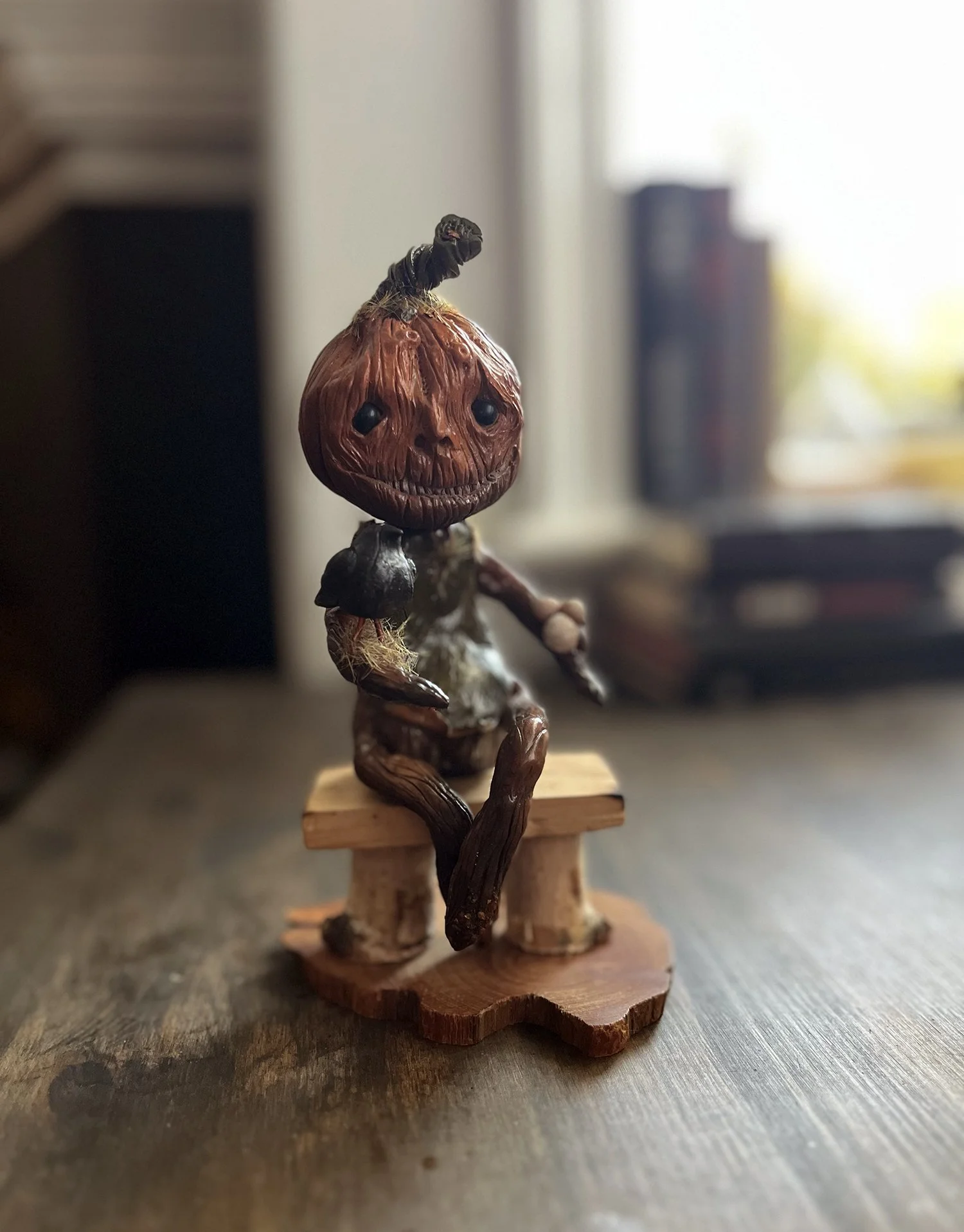 A handcrafted figurine of a pumpkin-headed creature with a stitched mouth, sitting on a small wooden chair, indoors with a blurred background.