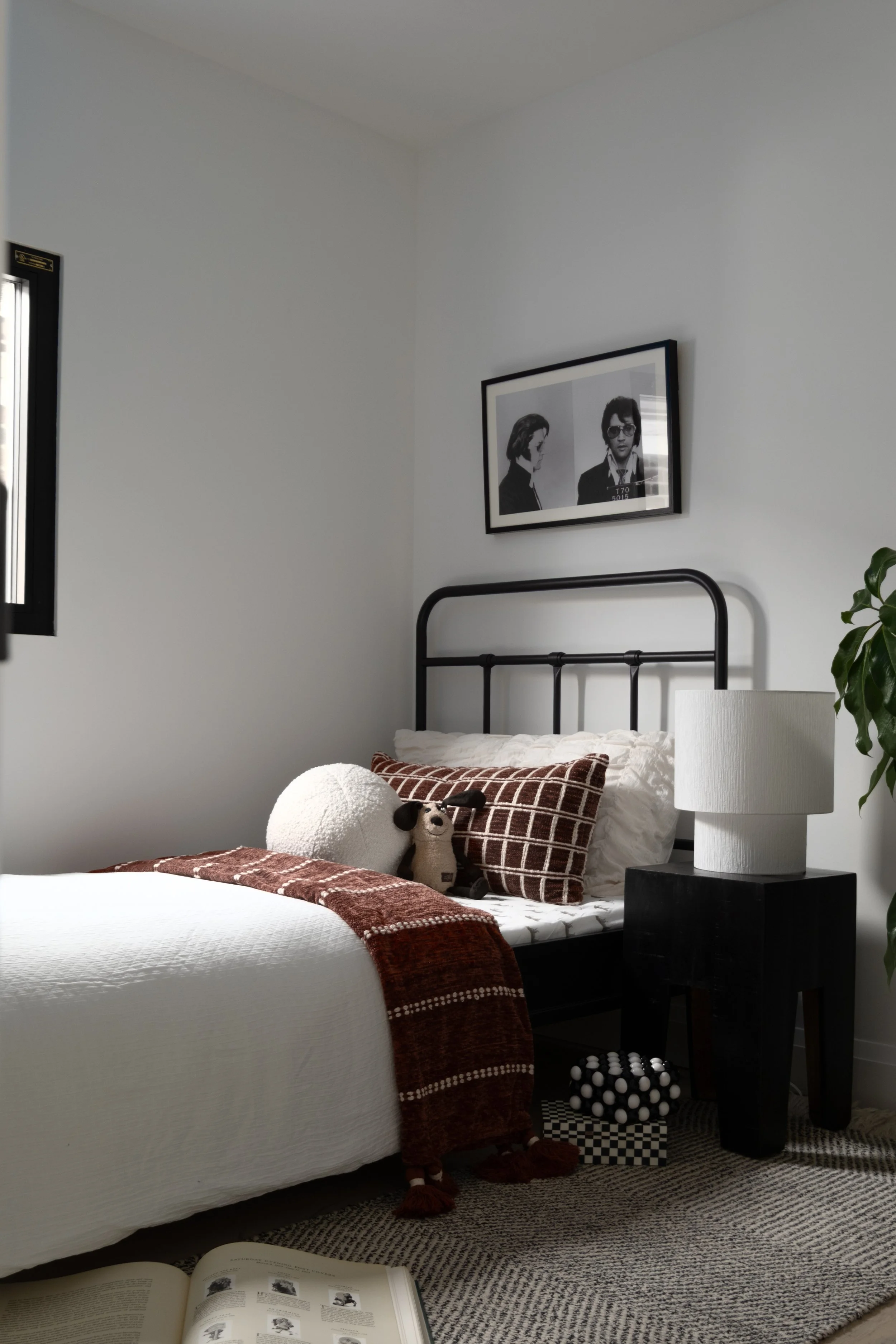 A bedroom with a bed featuring white bedding, a brown checkered pillow, a round plush white pillow with animal ears, and a small plush dog. There is a black side table with a large white lamp, a framed black-and-white photo on the wall, and a green p