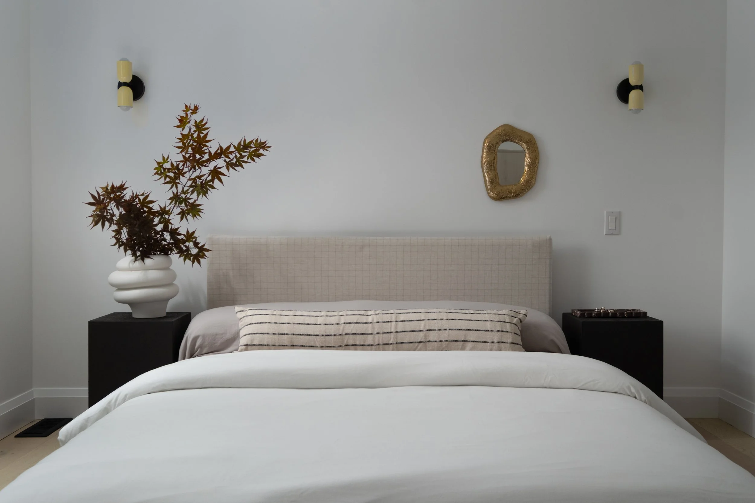 Minimalist bedroom with a bed, a beige headboard, black nightstands, a large white vase with red leaves, a small mirror, and wall sconces.