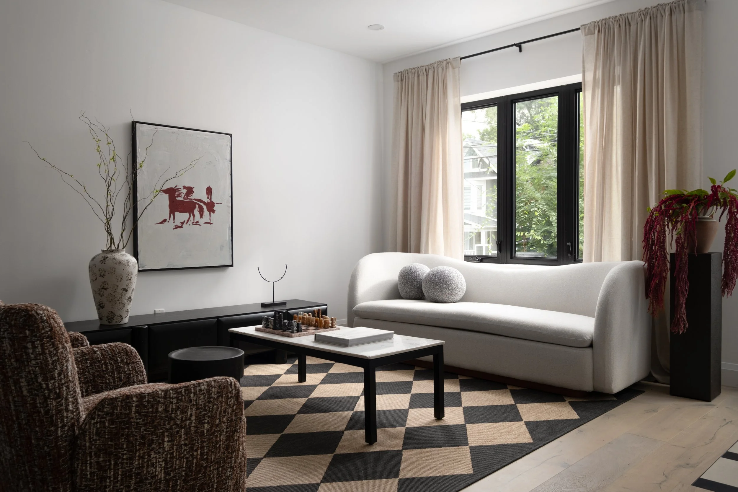 Living room with white sofa, black and white checkered rug, chessboard on coffee table, two patterned armchairs, large window with cream curtains, plants in vases, and art on the wall.