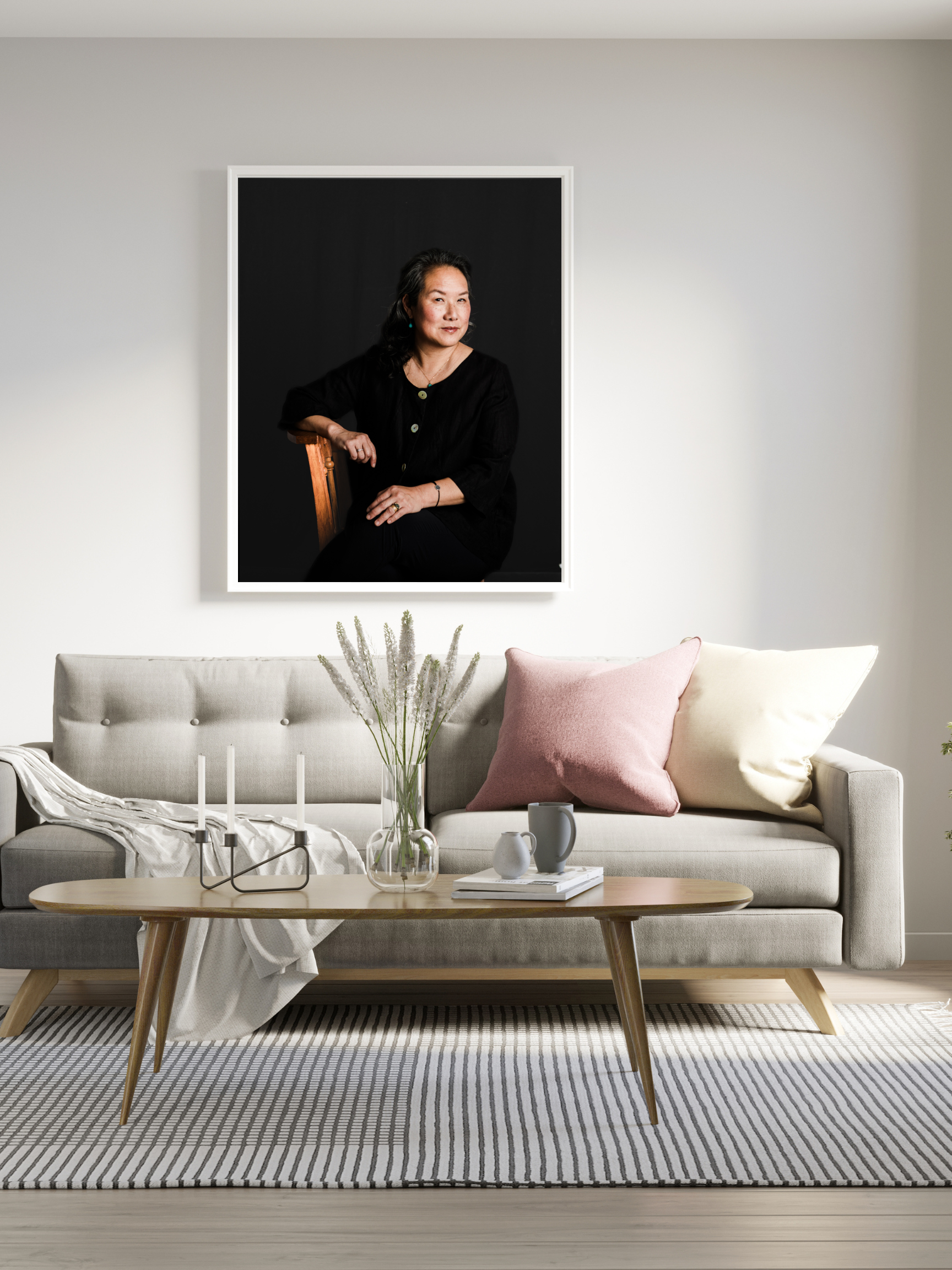 Living room with a light-colored sofa, pink and cream pillows, a wooden coffee table with vases and books, a patterned rug, and a large portrait of a woman hanging on a white wall.