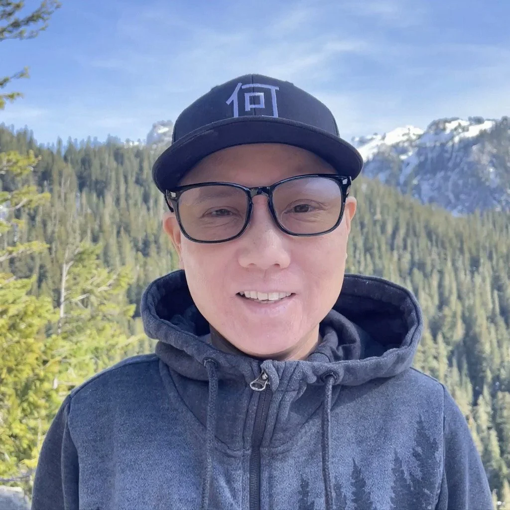 A woman outdoors in front of a mountain landscape with snow-capped peaks and a forest of evergreen trees, wearing a black cap, glasses, and a gray hoodie.