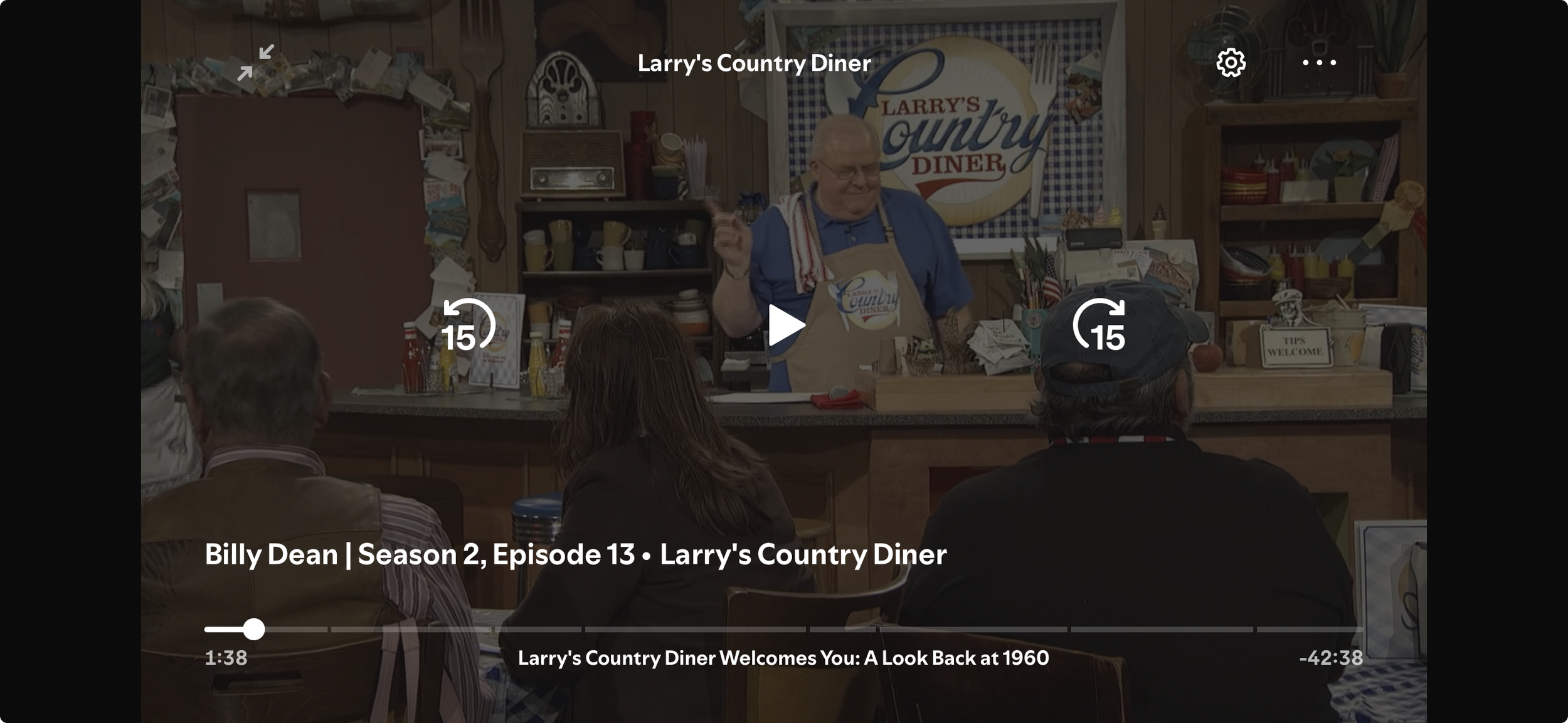 A scene from the TV show "Larry's Country Diner" featuring a host standing behind a counter, gesturing, with an audience seated in front. The background has a rustic wooden decor with a sign that reads "Larry's Country Diner."