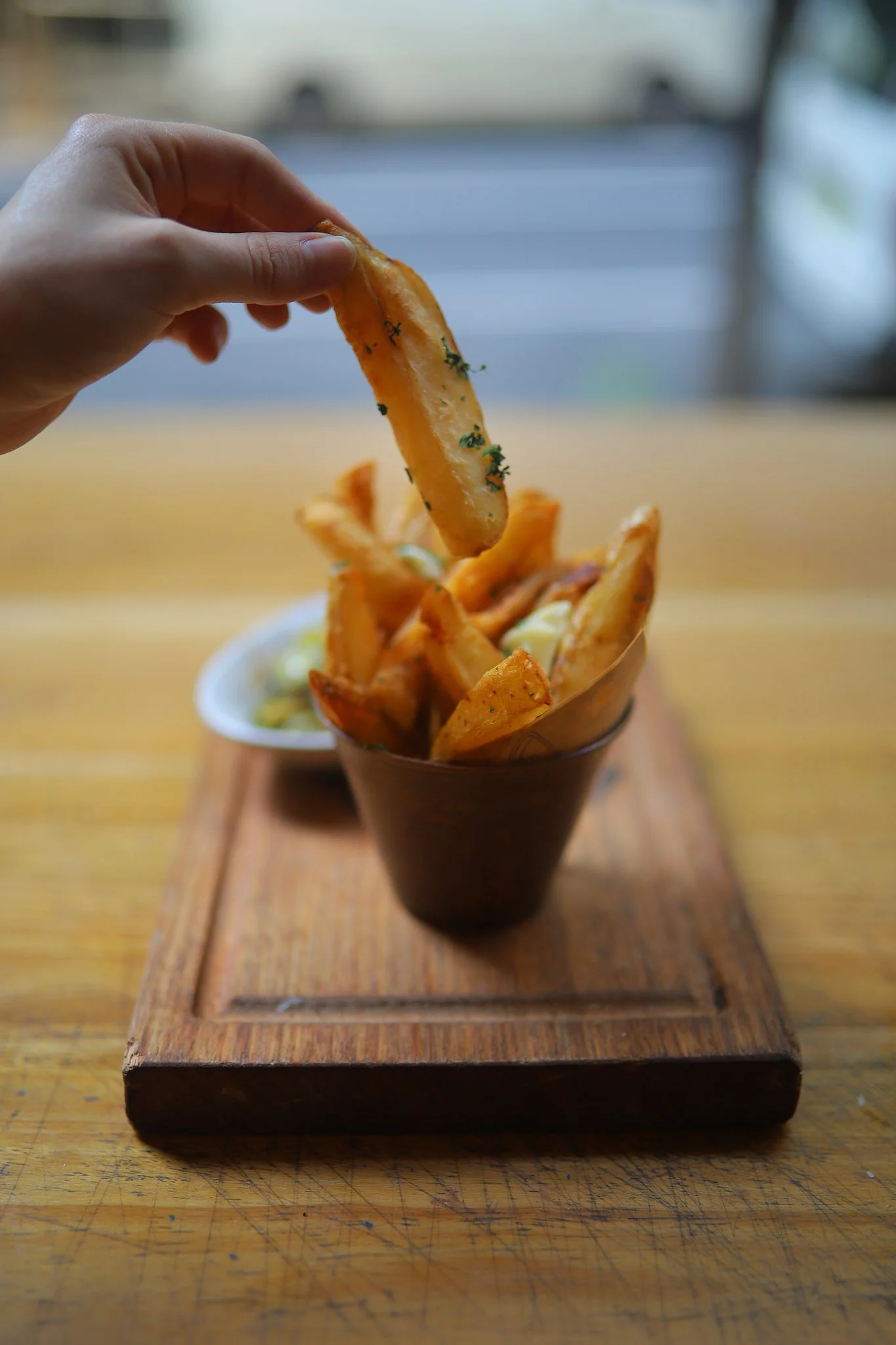 Hand holding a sweet potato fry above a cup of fries on a wooden serving tray.