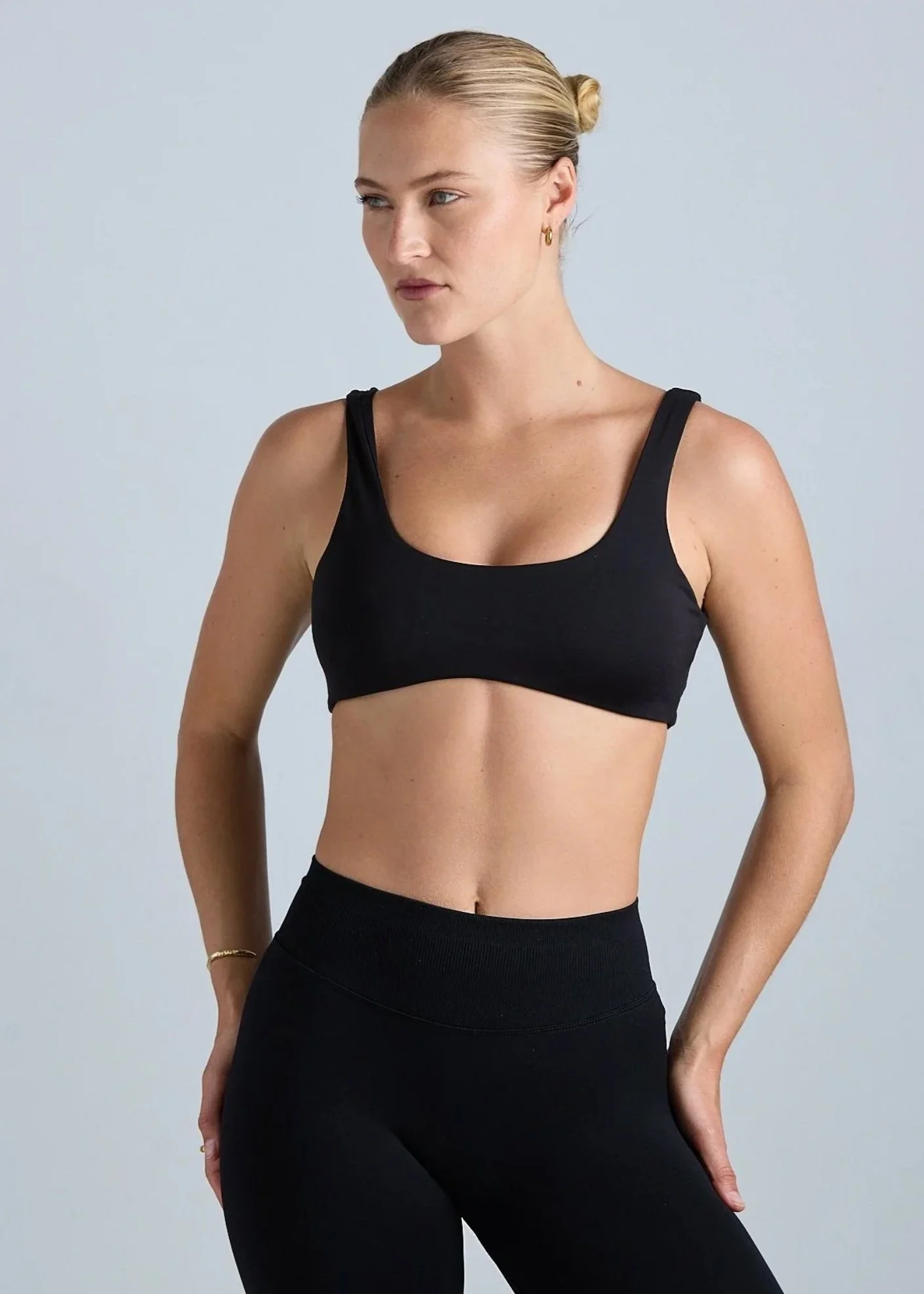 A woman with blonde hair tied back in a bun, wearing a black sports bra and black leggings, standing against a plain light gray background.