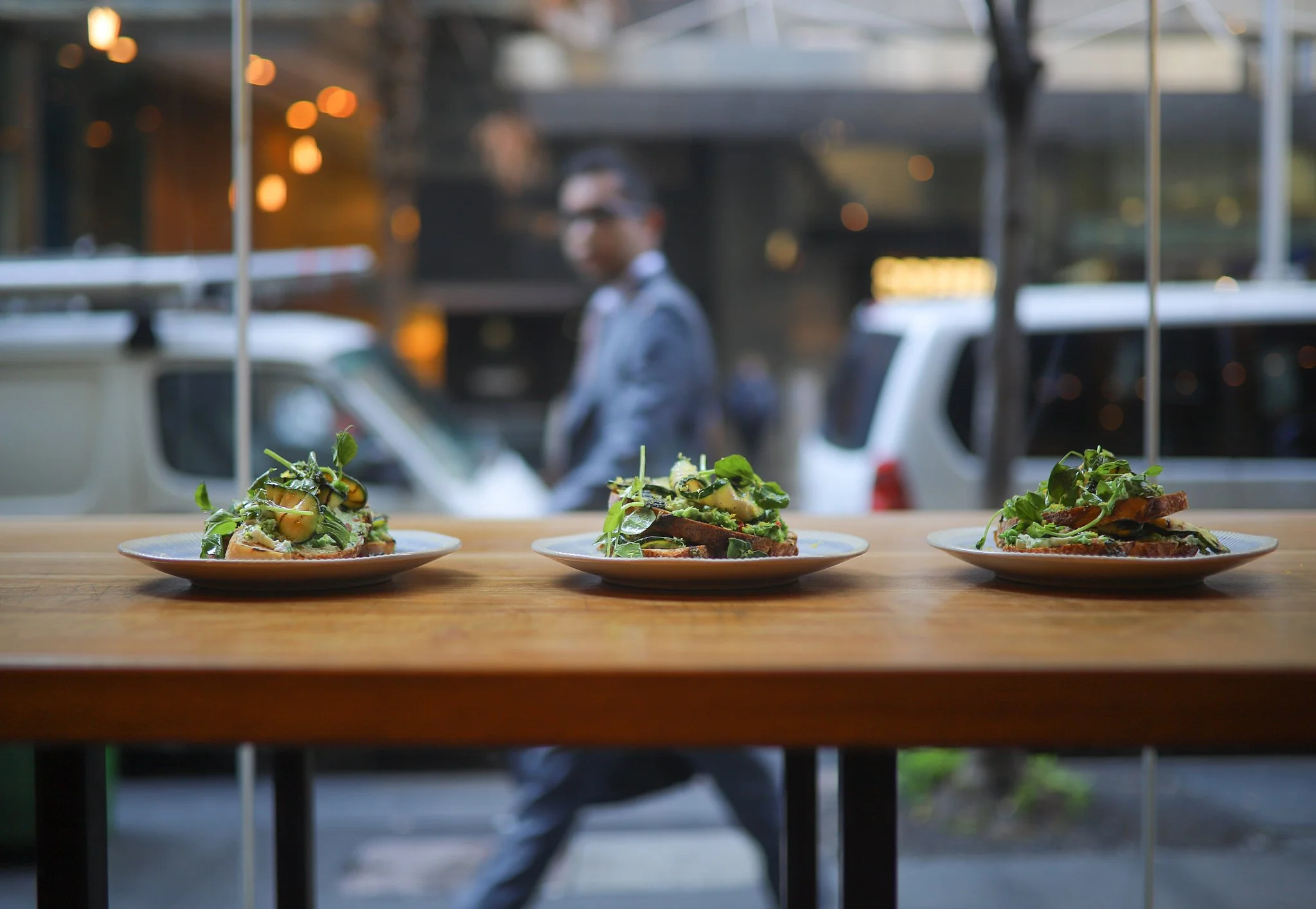 Three plates of open-faced sandwiches with greens and toppings sit on a wooden table inside a restaurant with a blurred view of a person walking outside.