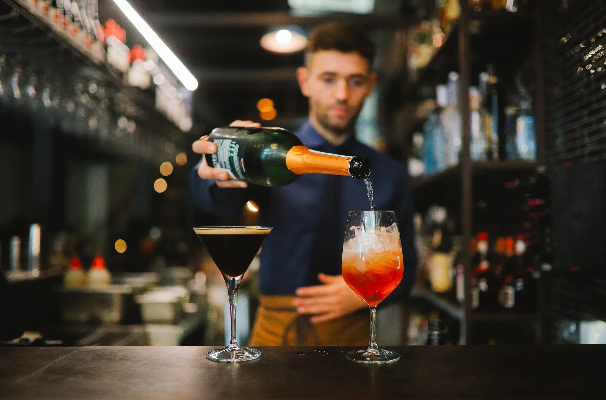 Bartender pouring a bottle into a glass with ice at a bar, with focused background lighting and shelves of bottles.
