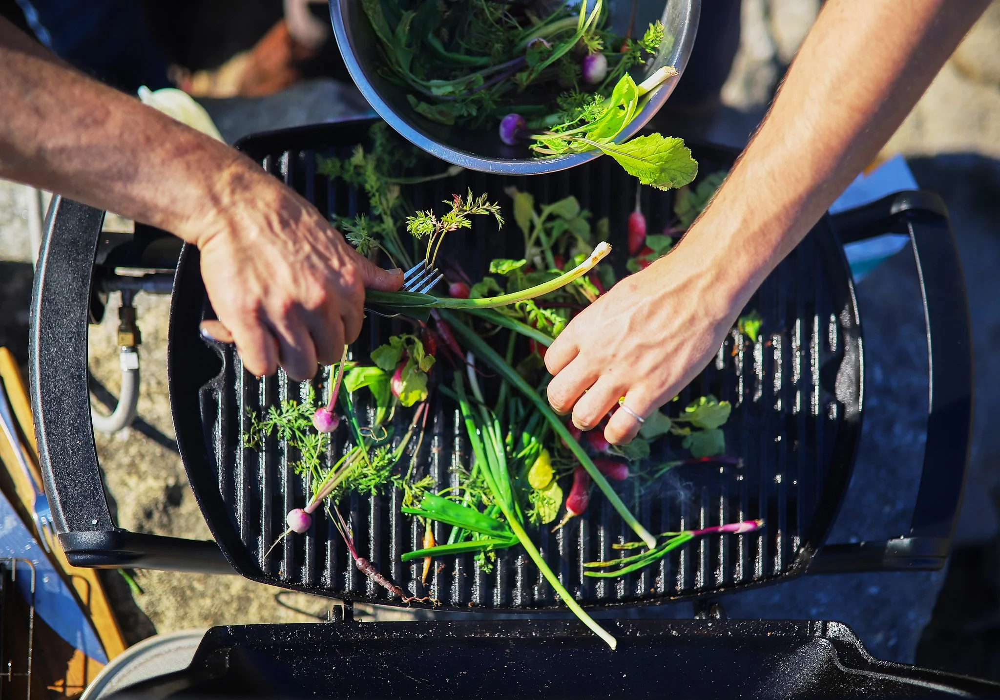 Hands harvesting radishes and greens from a black crate filled with vegetables outdoors on a sunny day.